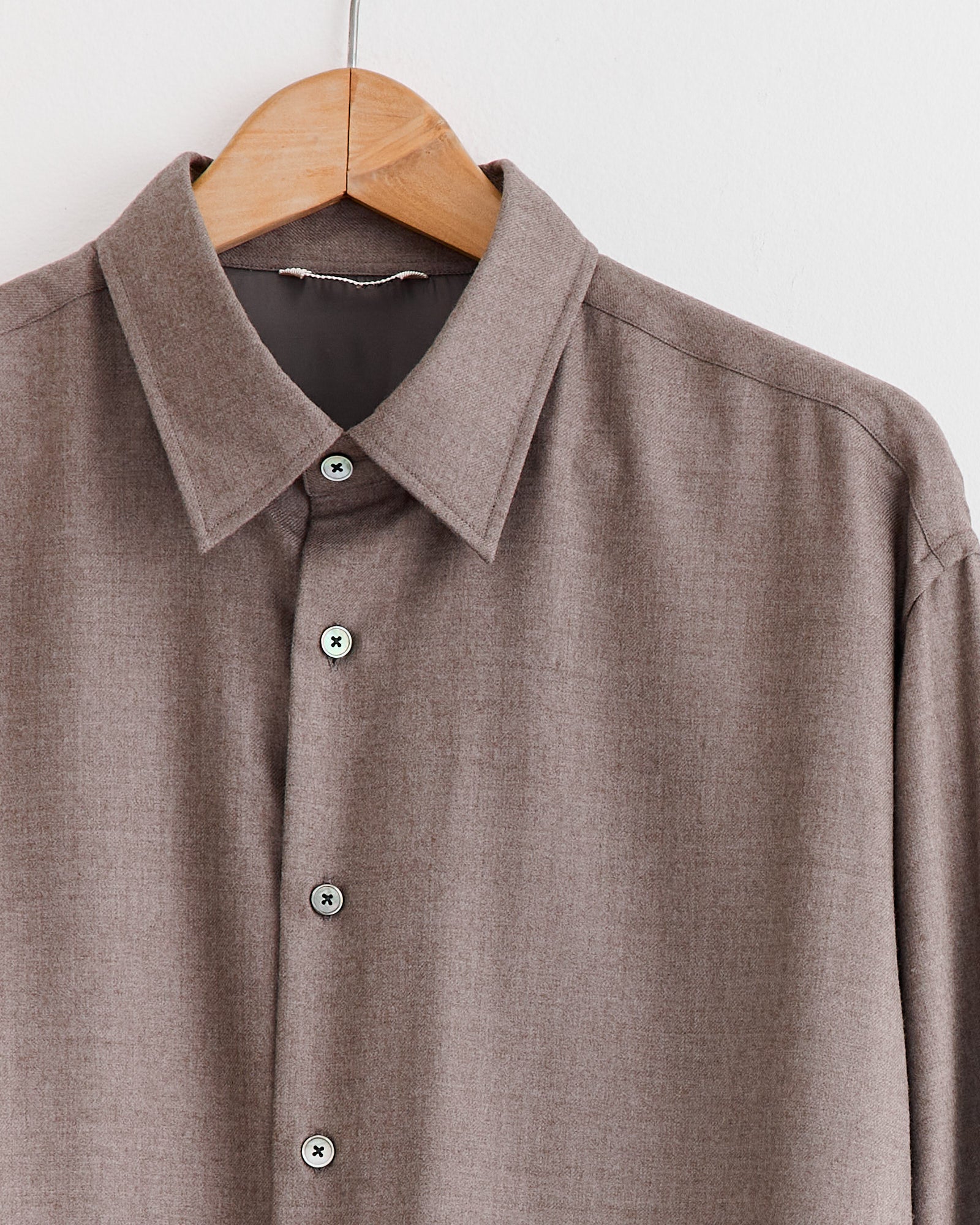 The Auralee Super Light Wool Shirt in Top Brown, featuring long sleeves, a pointed collar, and washable wool fabric, hangs on a wooden hanger against a white background.