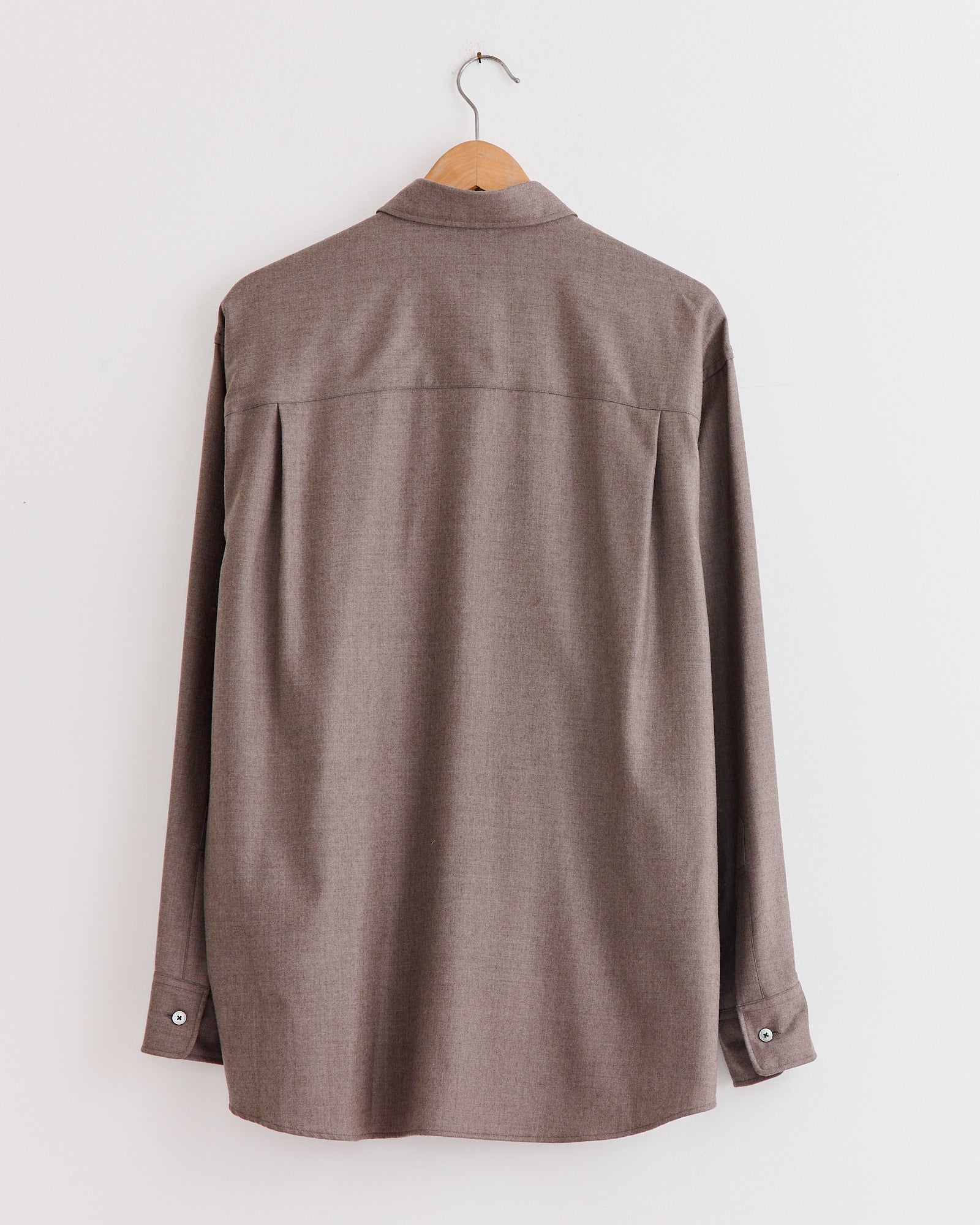 Auralee Super Light Wool Shirt in Top Brown with long sleeves hangs on a wooden hanger, photographed from the back against a plain white wall.