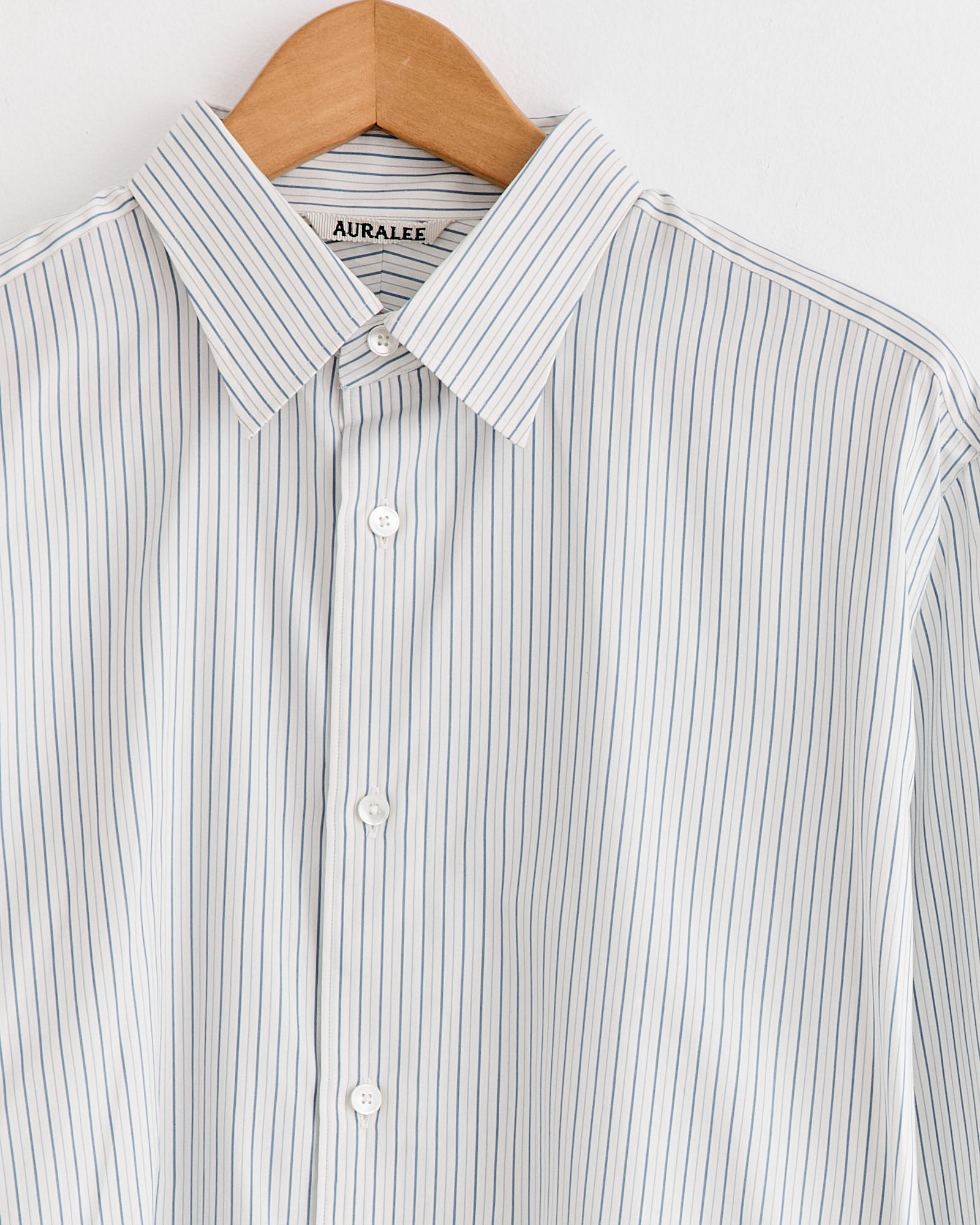 The Auralee Cotton Silk Stripe Shirt in White, made in Japan from fine cotton-silk blend, showcases light blue and white stripes, a classic collar, and white buttons—presented on a wooden hanger against a white background.