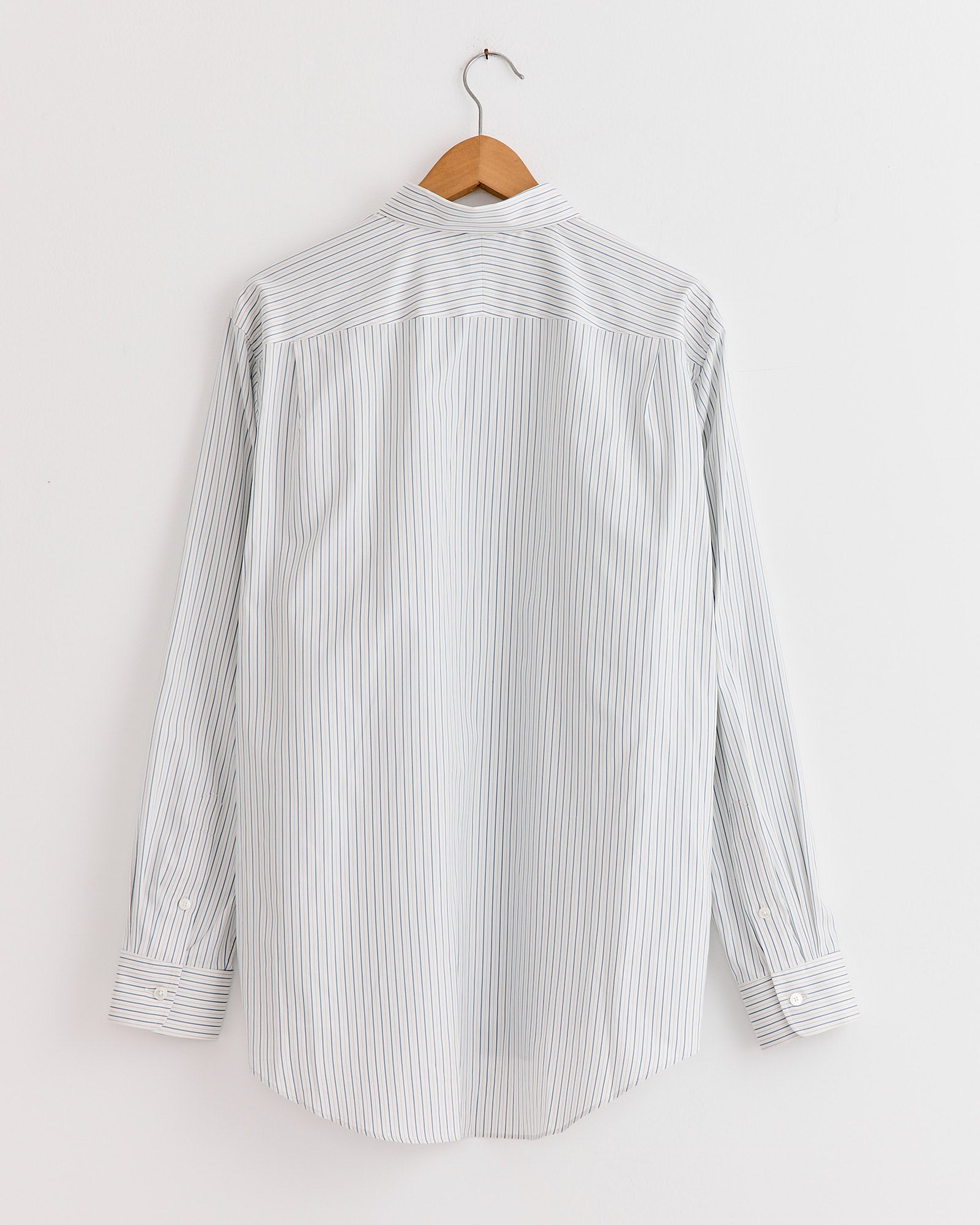 The Auralee Cotton Silk Stripe Shirt in White, crafted in Japan from premium Giza cotton, showcases fine blue stripes on crisp white fabric and is pictured from the back on a wooden hanger against a white wall.