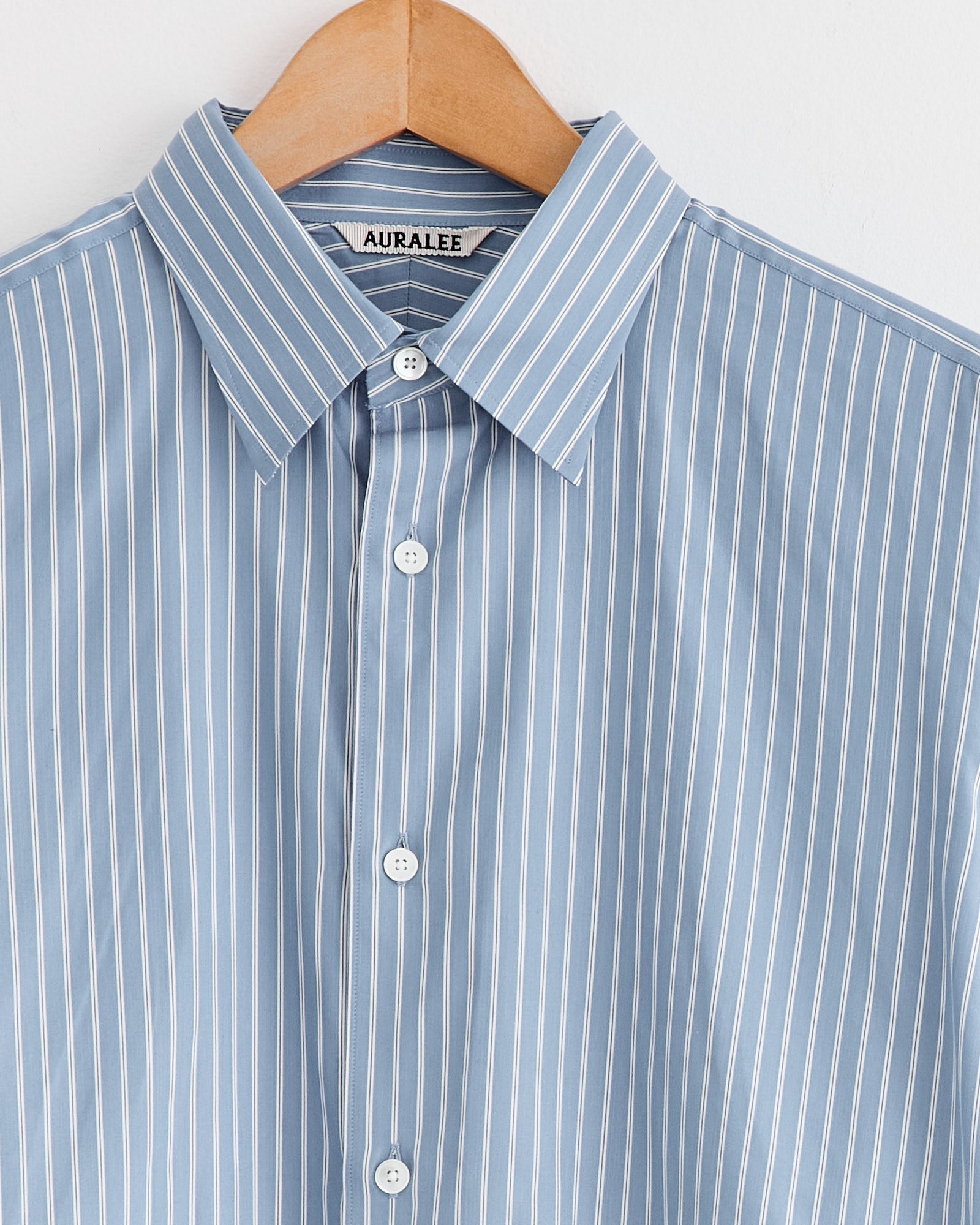A light blue Cotton Silk Stripe Shirt from Auralee, detailed with white vertical stripes, a pointed collar, and white buttons, hangs on a wooden hanger—showcasing the brand’s timeless luxury.