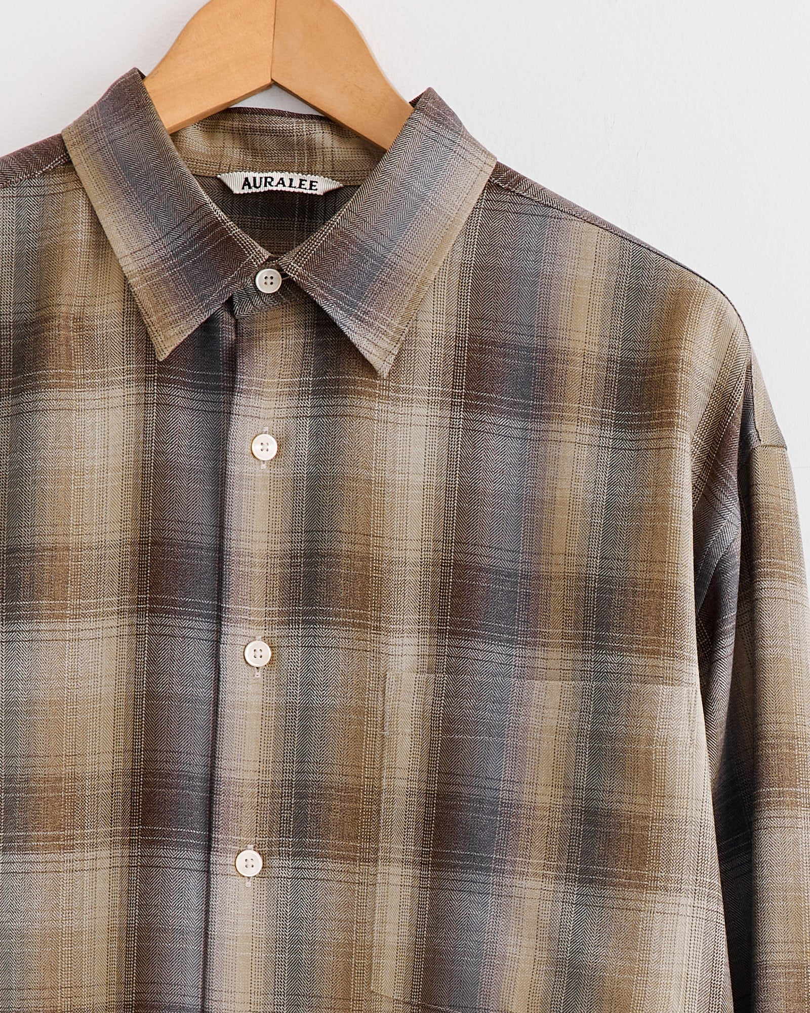 An Auralee Light Wool Check Shirt in brown and blue plaid, made in Japan, hangs on a wooden hanger against a white background. The label inside the collar reads “AURALEE.”.