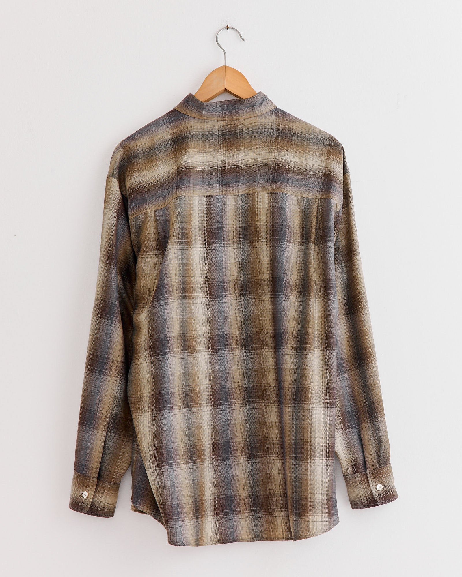 The Auralee Light Wool Check Shirt in Brown Blue, crafted in Japan from soft Super 120’s wool, hangs on a wooden hanger against a white wall, displayed from the back featuring brown, beige, and blue plaid.