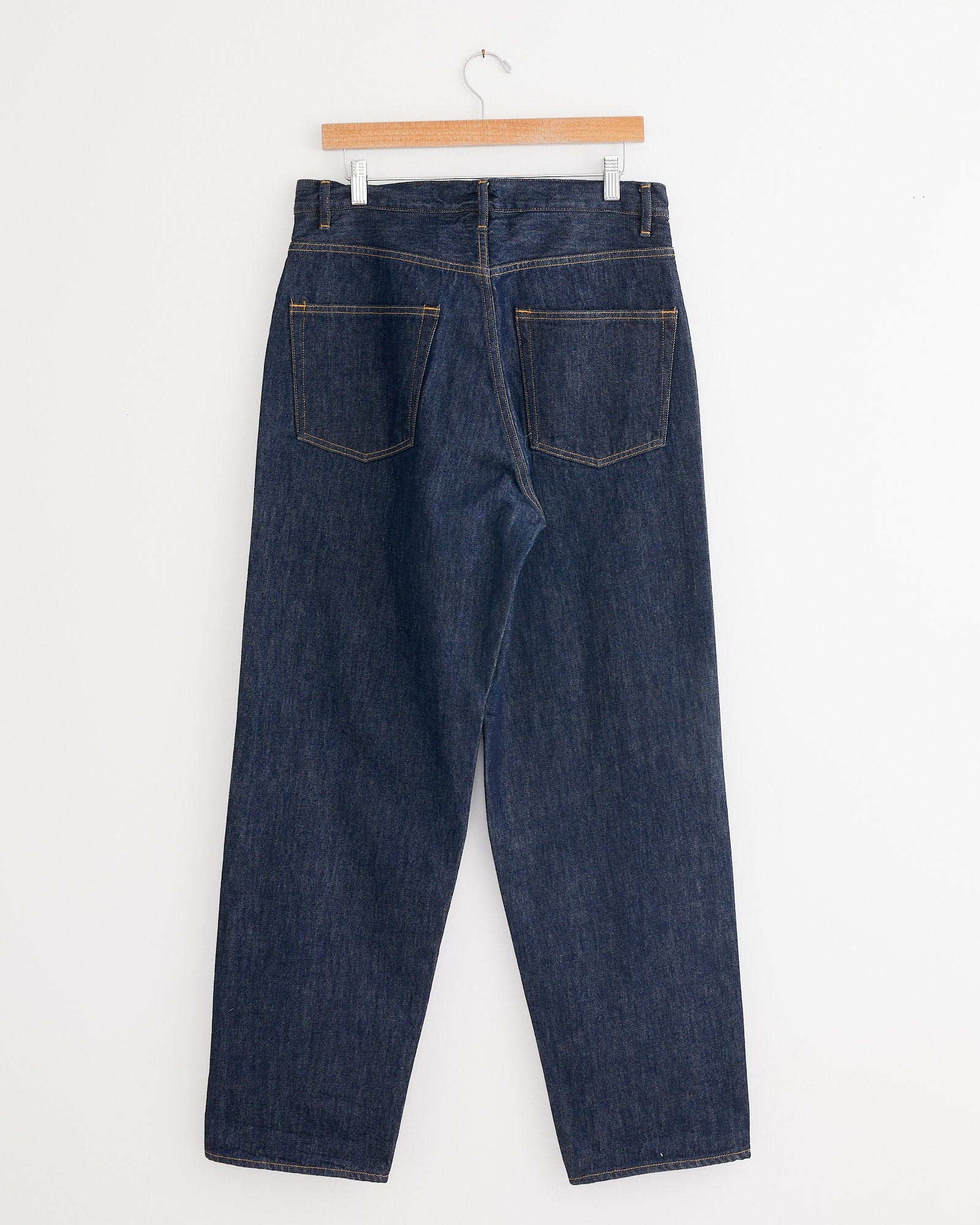 Auralee’s New Hard Twist Denim Pant in Indigo hangs on a wooden hanger against a white wall, shown from the back with straight legs and two large back pockets, crafted from premium Japanese raw denim.