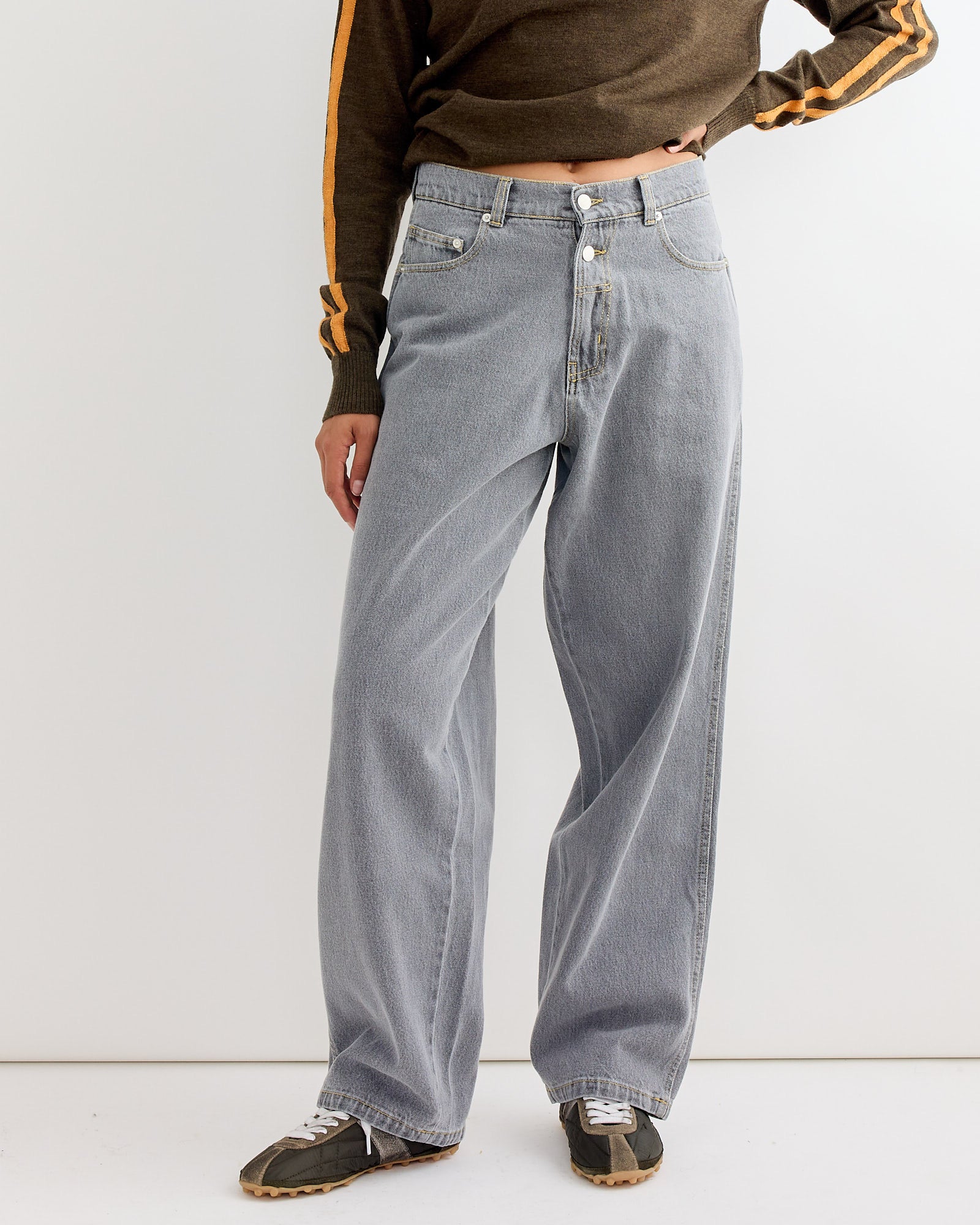 A person stands against a white background in Gimaguas Baggy Trouser in Grey, paired with a brown long-sleeve top with orange sleeve stripes and black sneakers with white laces, showcasing effortless style.