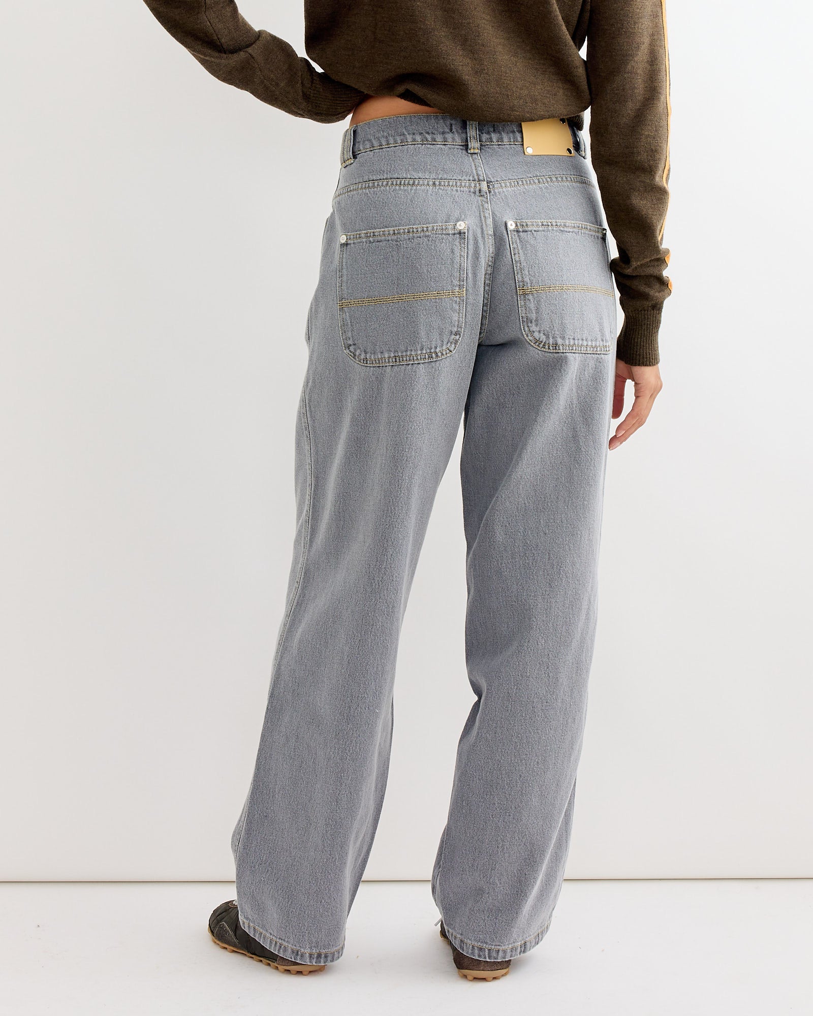 A person stands with their back to the camera, wearing the Gimaguas Baggy Trouser in Grey and a brown long-sleeve top, paired with brown shoes.