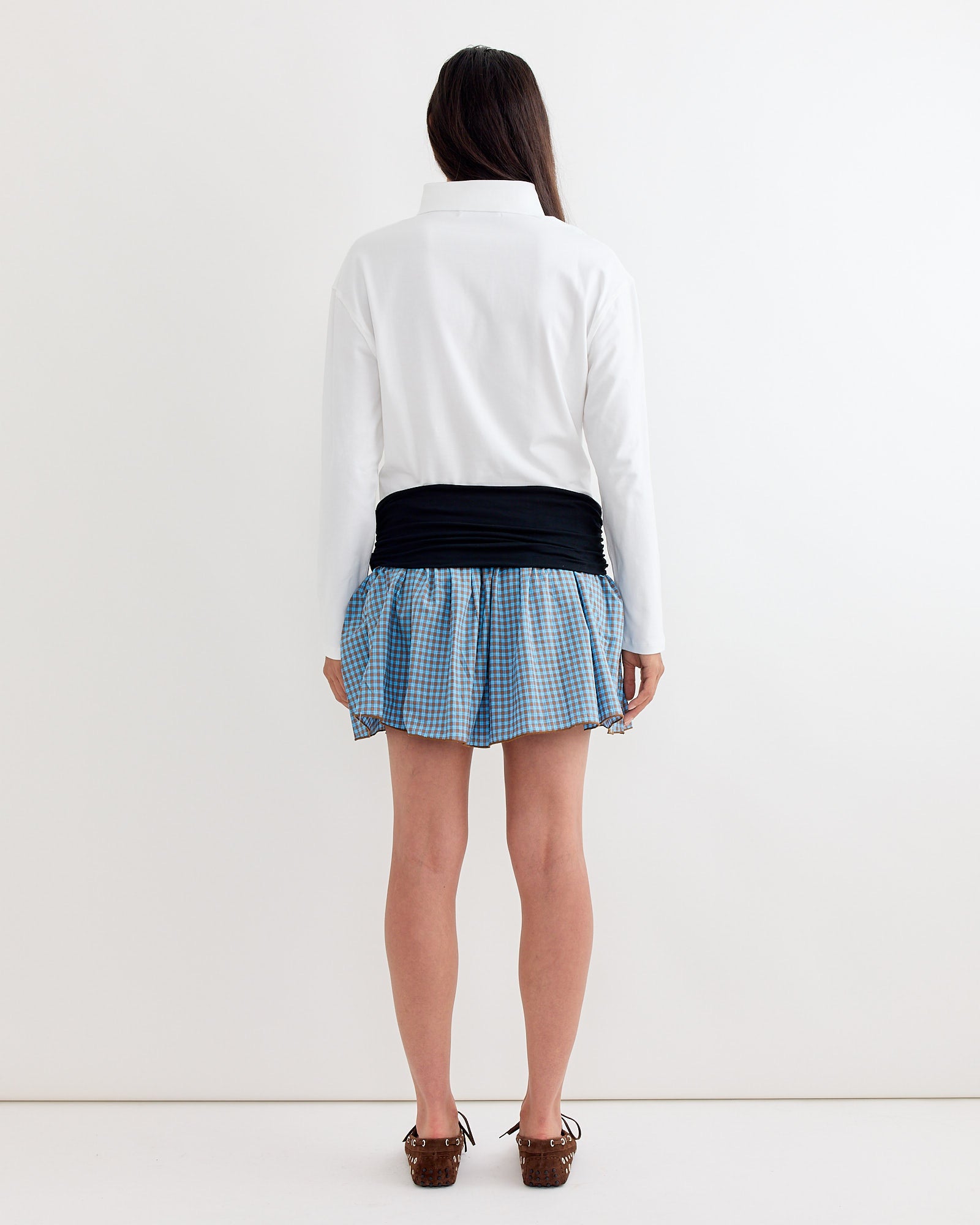 A person stands facing a white wall, wearing the Chico Mini Dress in White by Gimaguas. Their straight dark hair falls down their back.