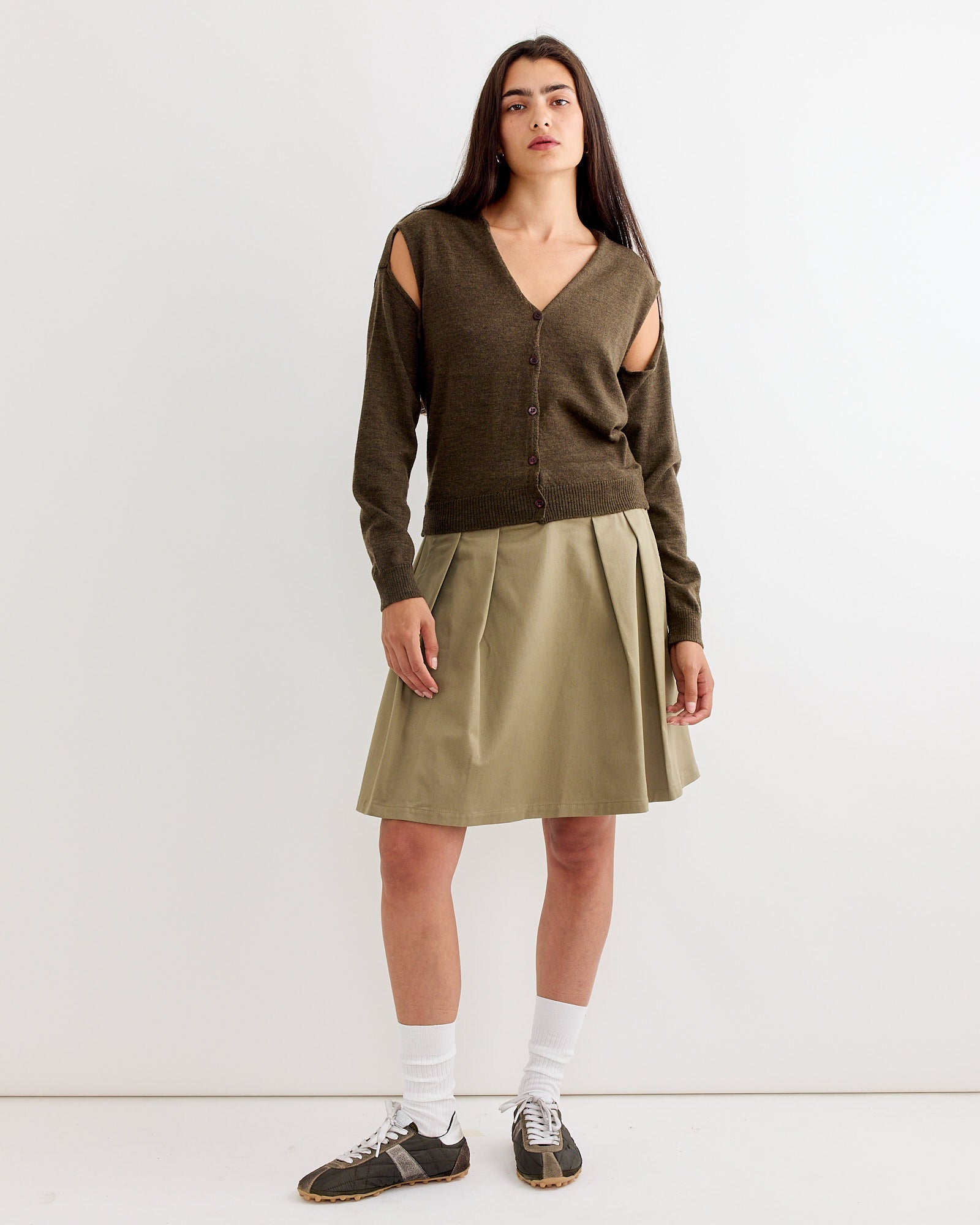 A woman with long dark hair wears the Gimaguas Coucou Cardigan in Brown, paired with a light olive pleated skirt, white socks, and black sneakers, standing against a plain white background.