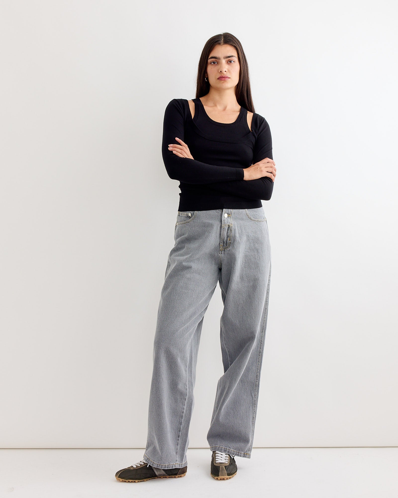 A woman with long dark hair stands against a white background, wearing the Gimaguas Etoile Jumper in Black, paired with loose-fitting light blue jeans and black sneakers. Her arms are crossed and she has a neutral expression.