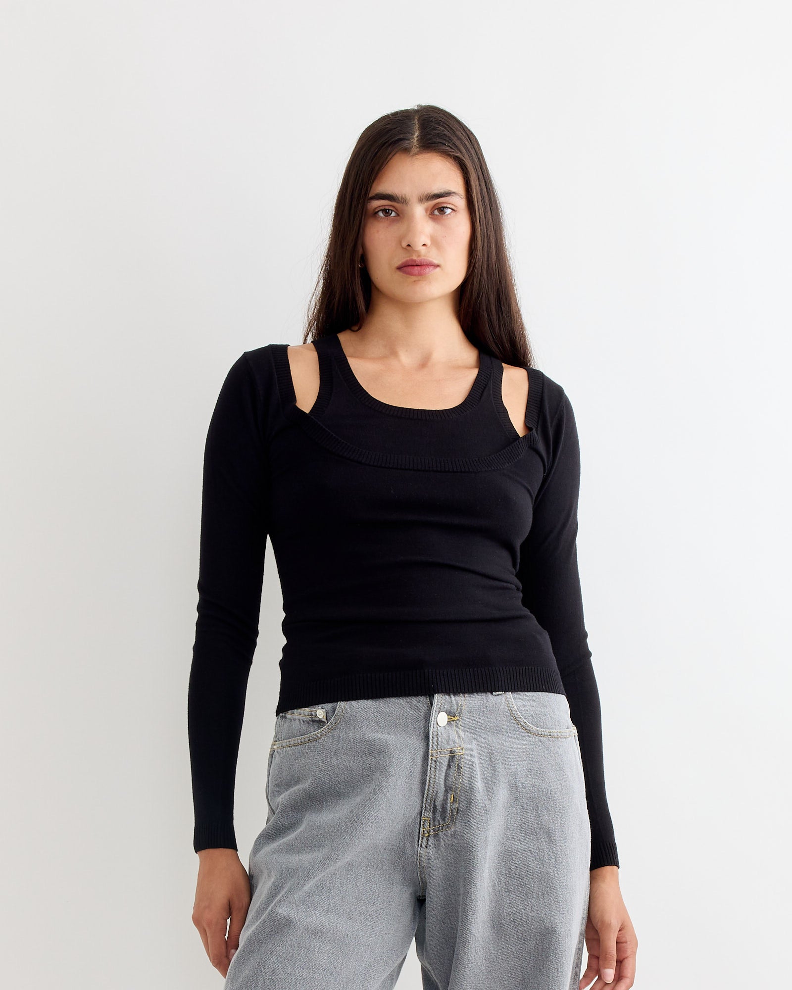 A woman with long dark hair wears the Gimaguas Etoile Jumper in Black, featuring cut-out shoulders, paired with light blue jeans, standing against a plain white background.