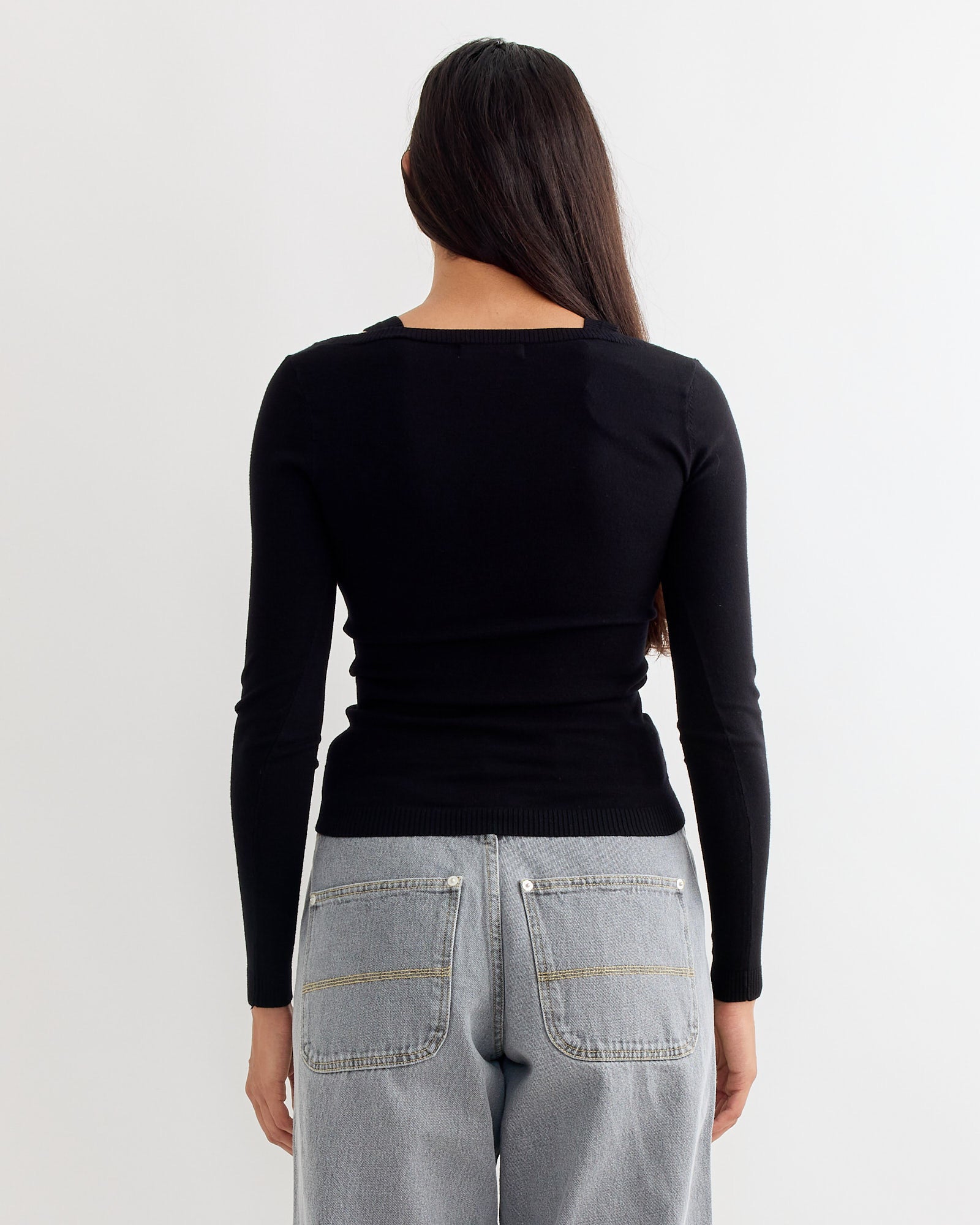 A person with long dark hair wears the Gimaguas Etoile Jumper in Black, paired with light gray jeans with large back pockets, standing against a plain white background facing away from the camera.