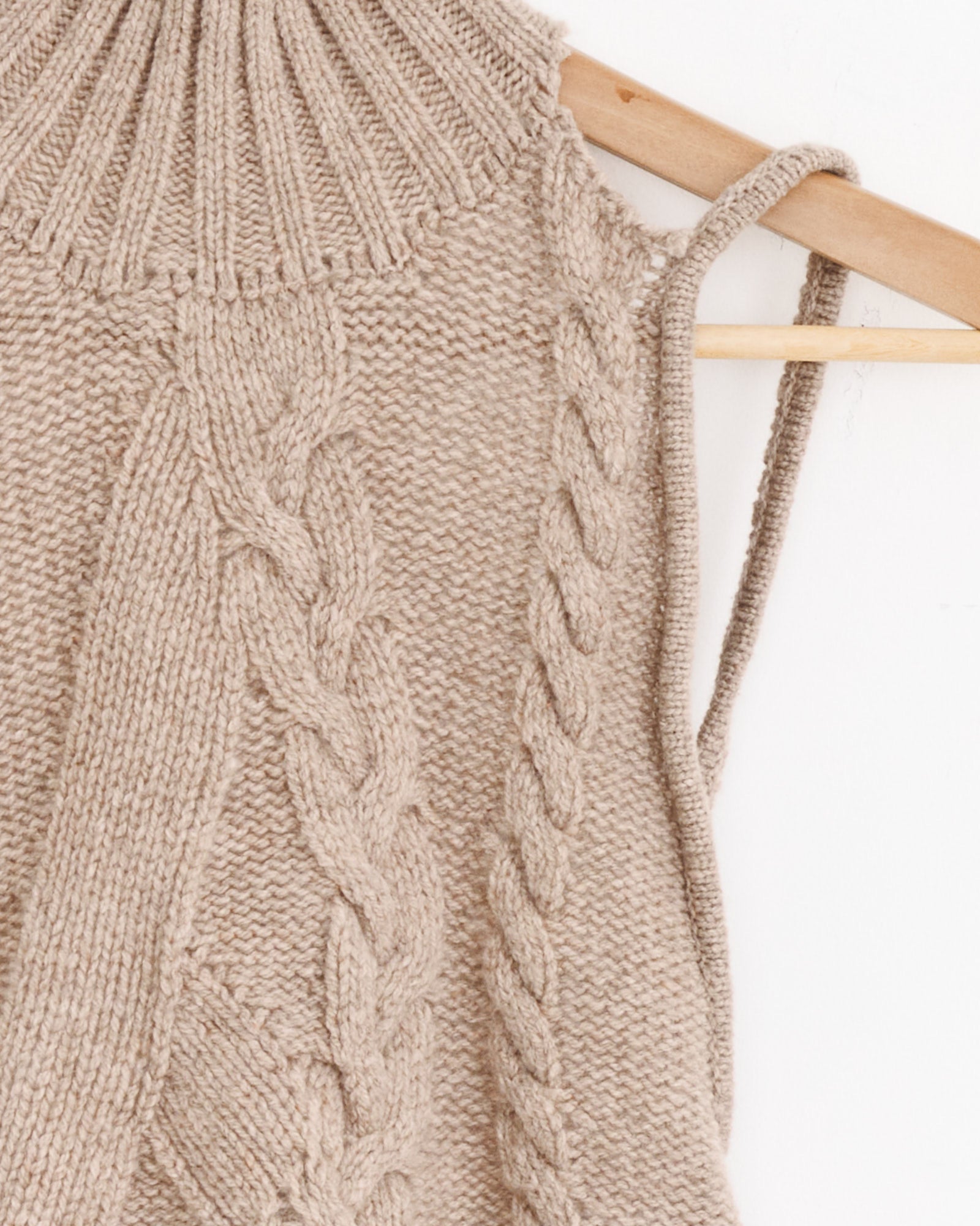 Close-up of the Gimaguas Julita Top in Beige, a sleeveless knit with cable patterns, ribbed high neckline, and a thin shoulder strap, crafted from extrafine wool. The piece is displayed on a wooden hanger against a white background.