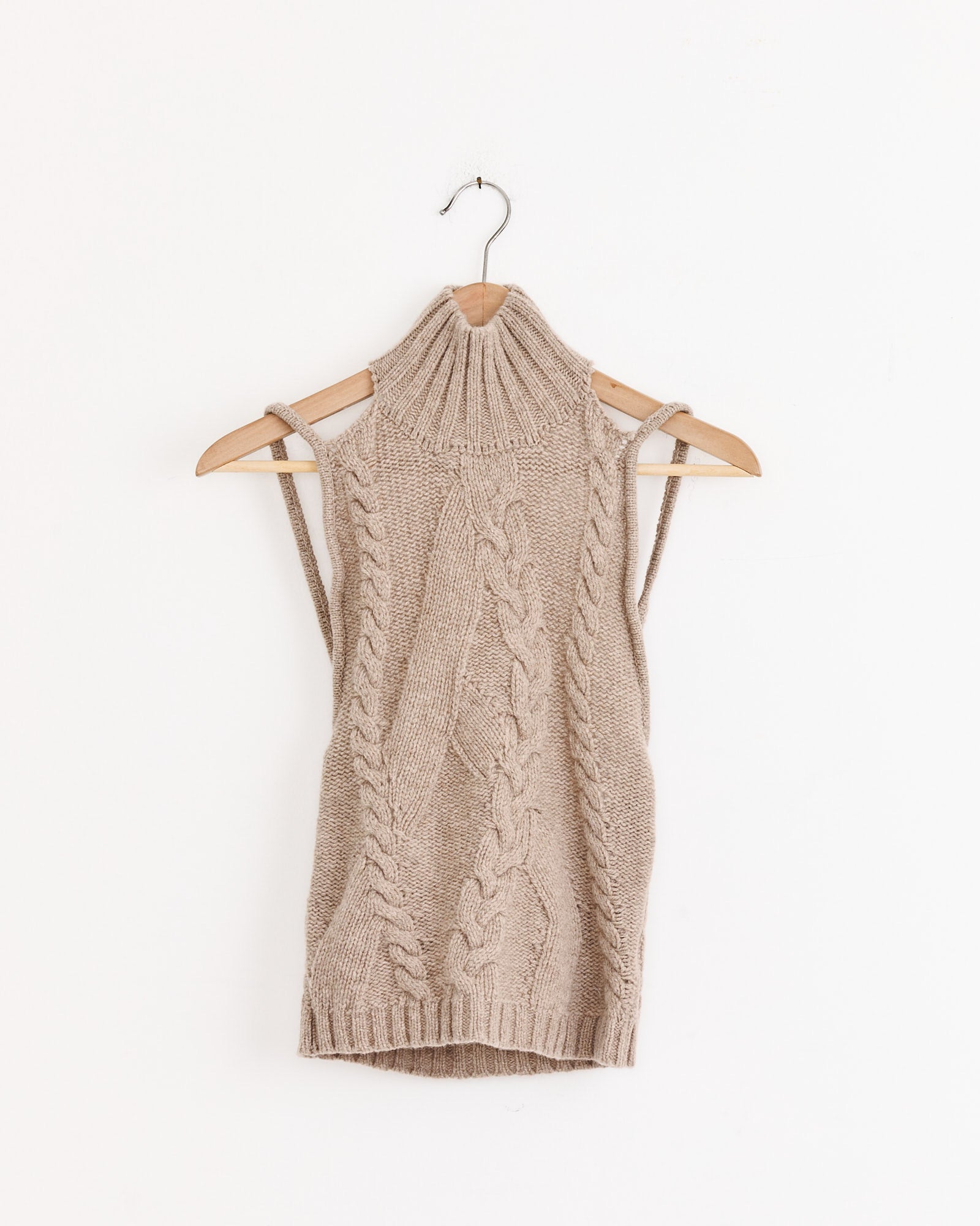The Julita Top in Beige by Gimaguas, a sleeveless extrafine wool knit with a cable pattern and ribbed turtleneck, hangs on a wooden hanger against a plain white background.
