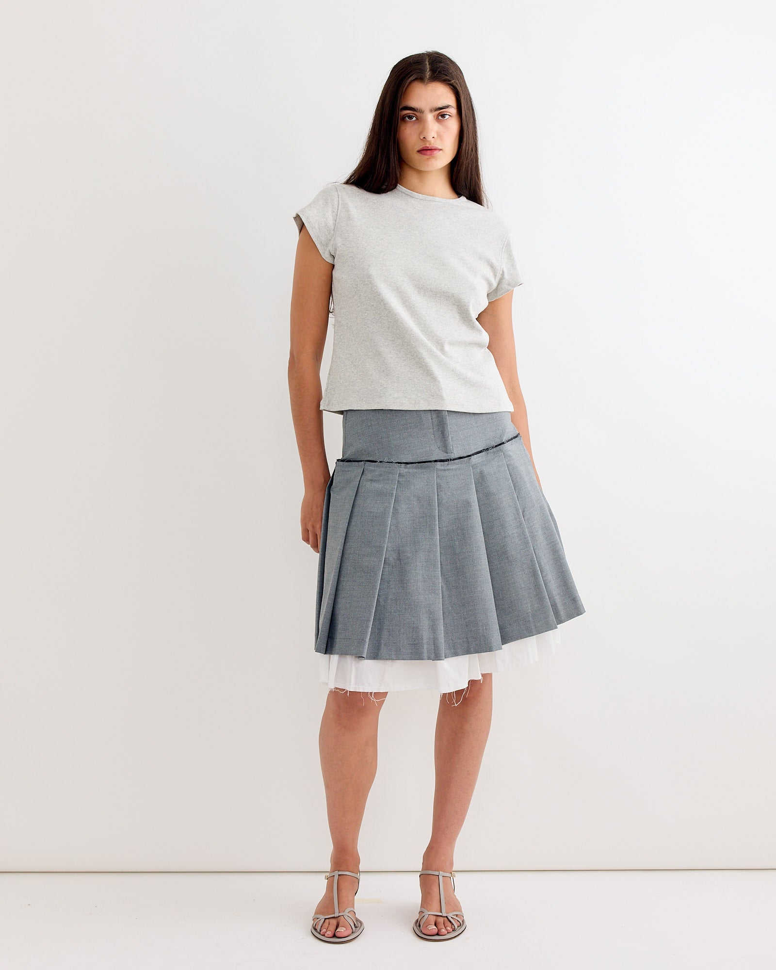 A woman with long brown hair wears the Gimaguas Manna Top in Grey, a pleated gray knee-length skirt with a white tulle layer, and silver strappy sandals, standing against a plain white background.