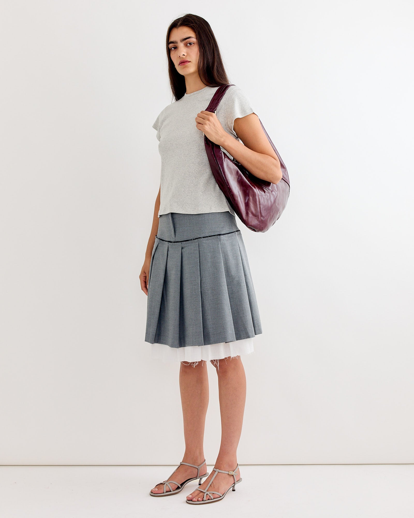 A woman wears a light gray short-sleeve top, the Gimaguas Tailored Midi Skirt in Grey, silver strappy heels, and carries a large maroon shoulder bag while standing against a plain white background.