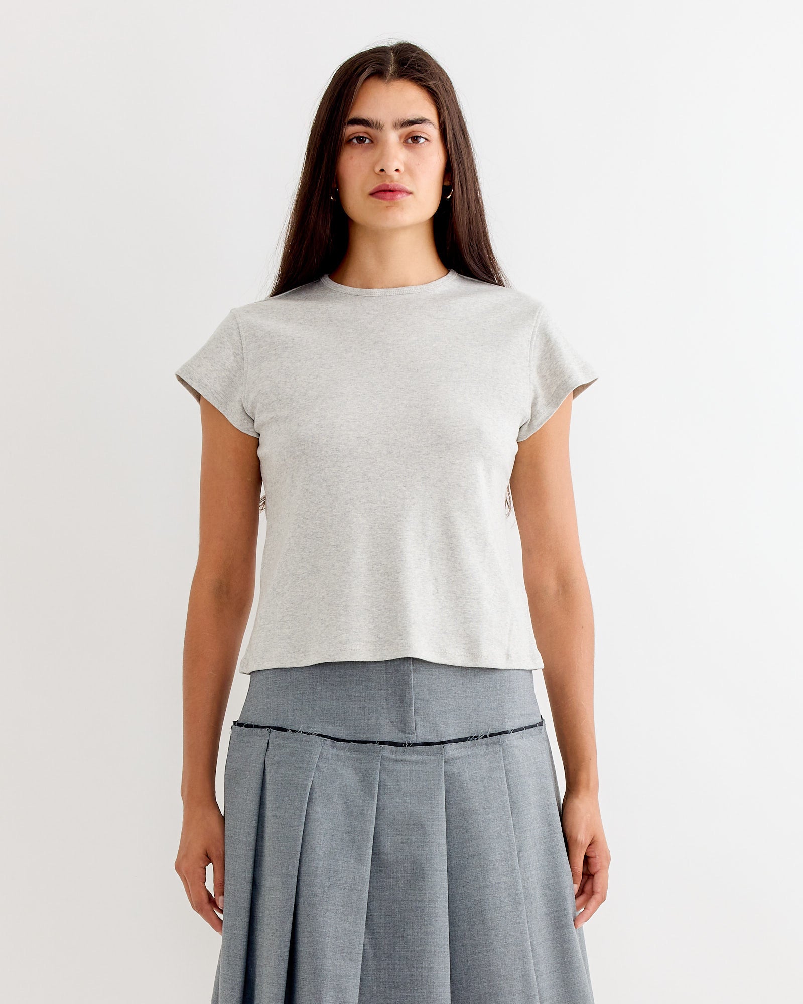 A woman with long dark hair wears the Gimaguas Manna Top in Grey, paired with a gray pleated skirt. She stands against a plain white background, facing forward with a neutral expression.