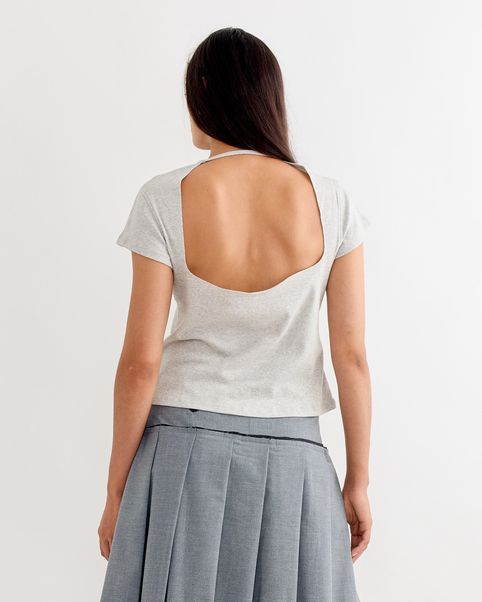 A woman with long dark hair, wearing the Gimaguas Manna Top in Grey with an open back and a pleated gray skirt, stands facing away against a plain white background.