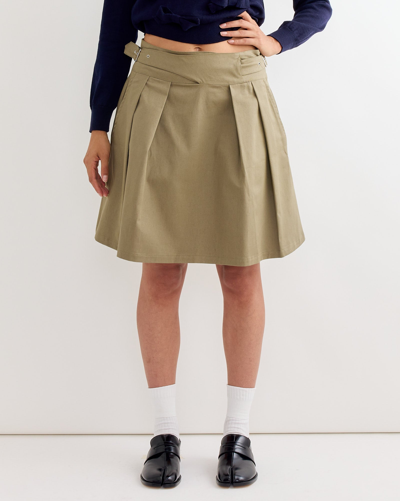 A person wearing the Gimaguas Martin Midi Skirt in Light Khaki with pleats, a navy-blue top, white socks, and black shoes stands against a plain white background, one hand on their hip.