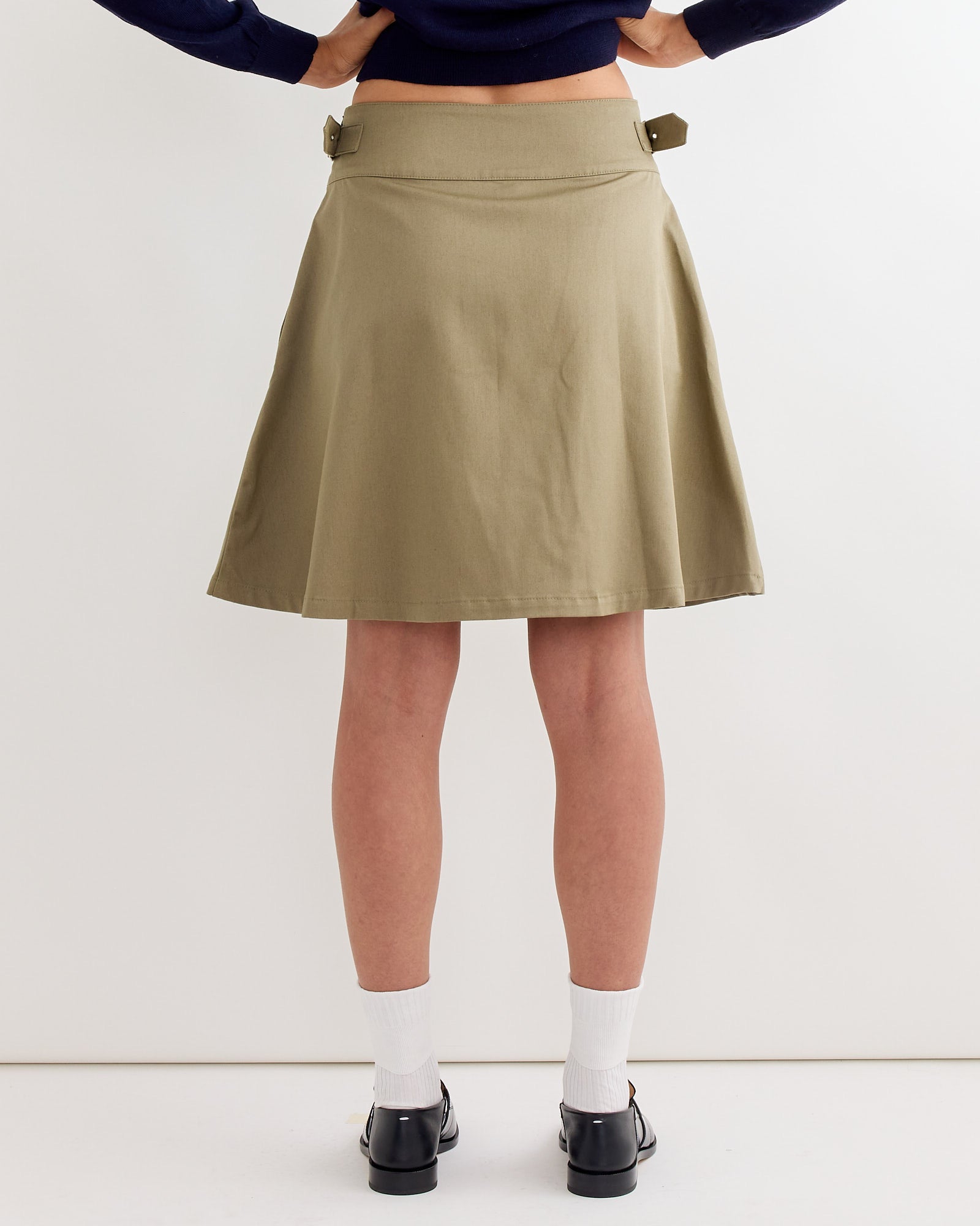 A model is shown from the back against a plain white background wearing the Gimaguas Martin Midi Skirt in Light Khaki, paired with white socks and black shoes.