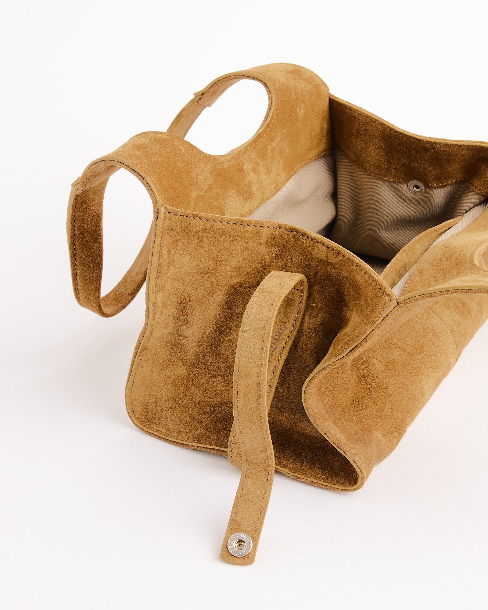 The Gimaguas Mini Maria Suede Bag is a tan shoulder bag with round handles, an open top, beige lining, and a snap button strap closure, shown against a white background.