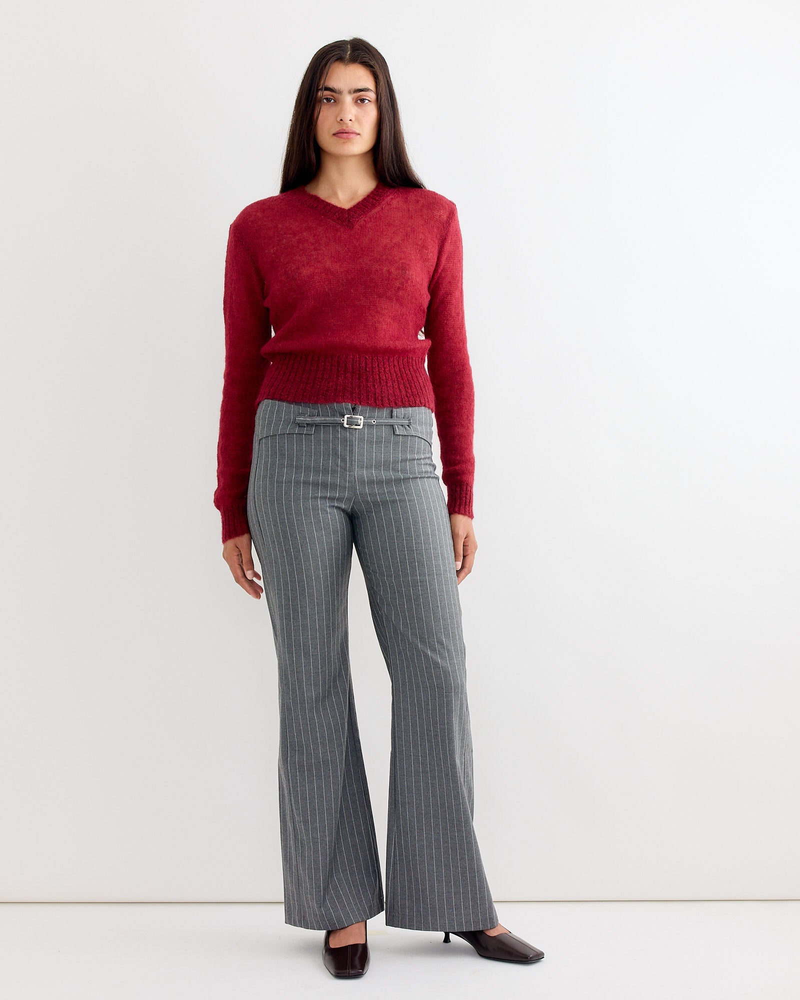 A woman with long dark hair stands against a white background, wearing the Gimaguas Mum Jumper in Burgundy, gray pinstriped belted pants, and dark heels. She has a neutral expression with her arms relaxed by her sides.