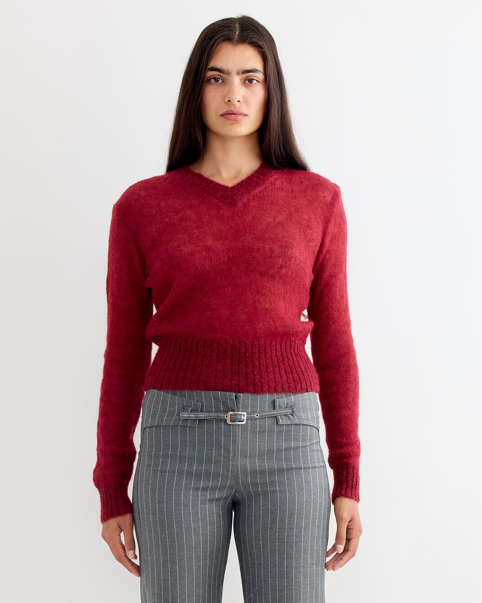 A person with long dark hair wears the Gimaguas Mum Jumper in Burgundy and gray pinstriped belted pants, standing against a plain white background.