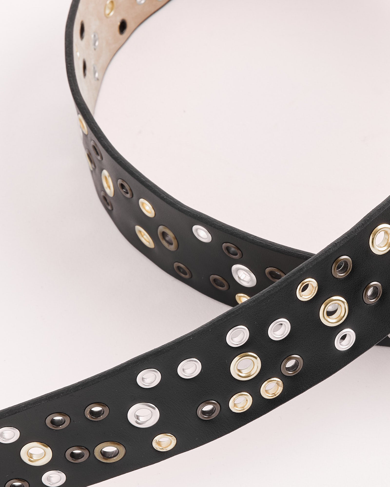 The Gimaguas Rufus Belt in Black features multiple rows of circular metal eyelets in silver, gold, and black, shown on a light background. For sizing and fit help, connect with us through our online chat.