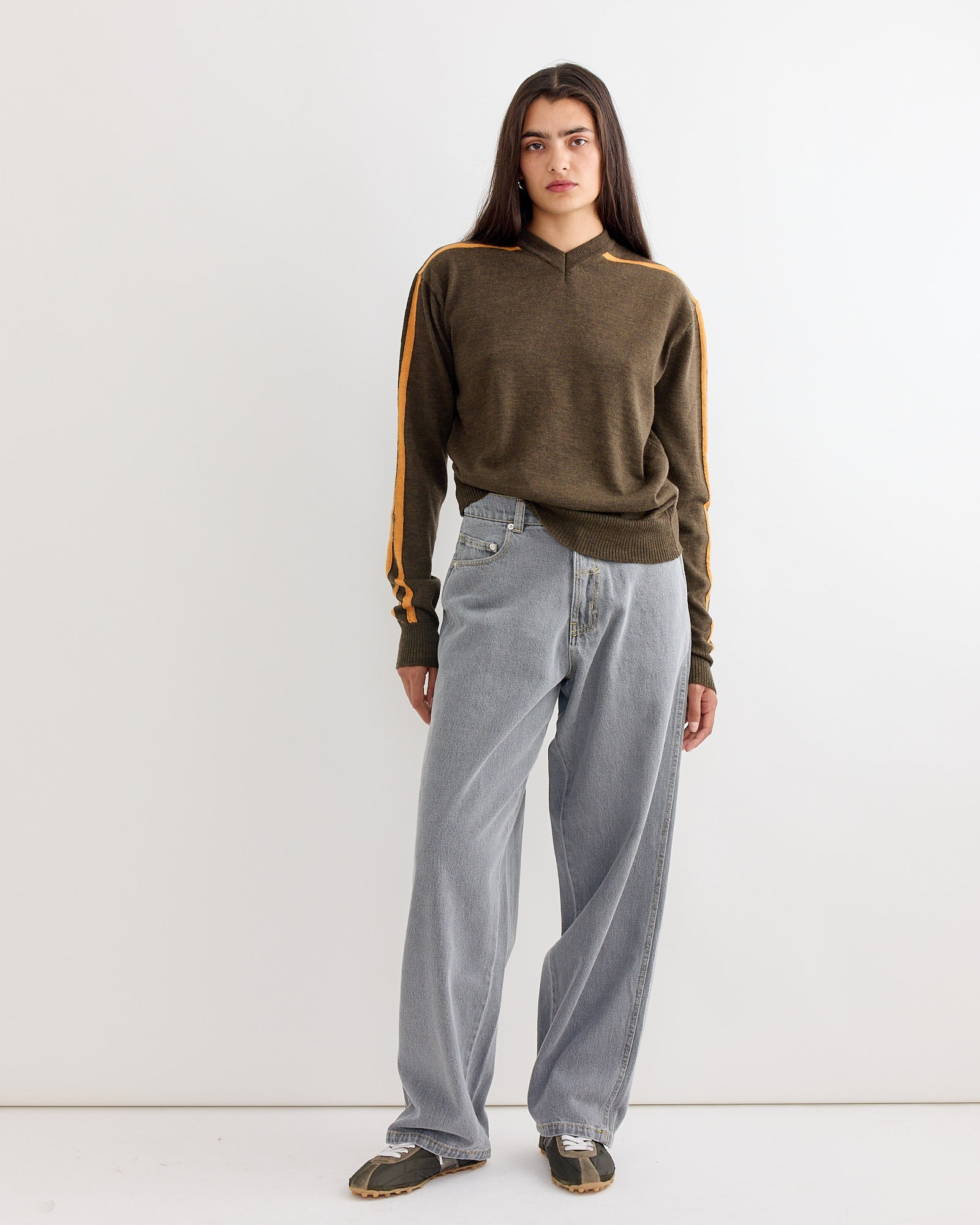 A woman with long dark hair models the Gimaguas Sport Jumper in Brown, featuring orange sleeve stripes, paired with light blue loose jeans and green sneakers, standing against a plain white background.