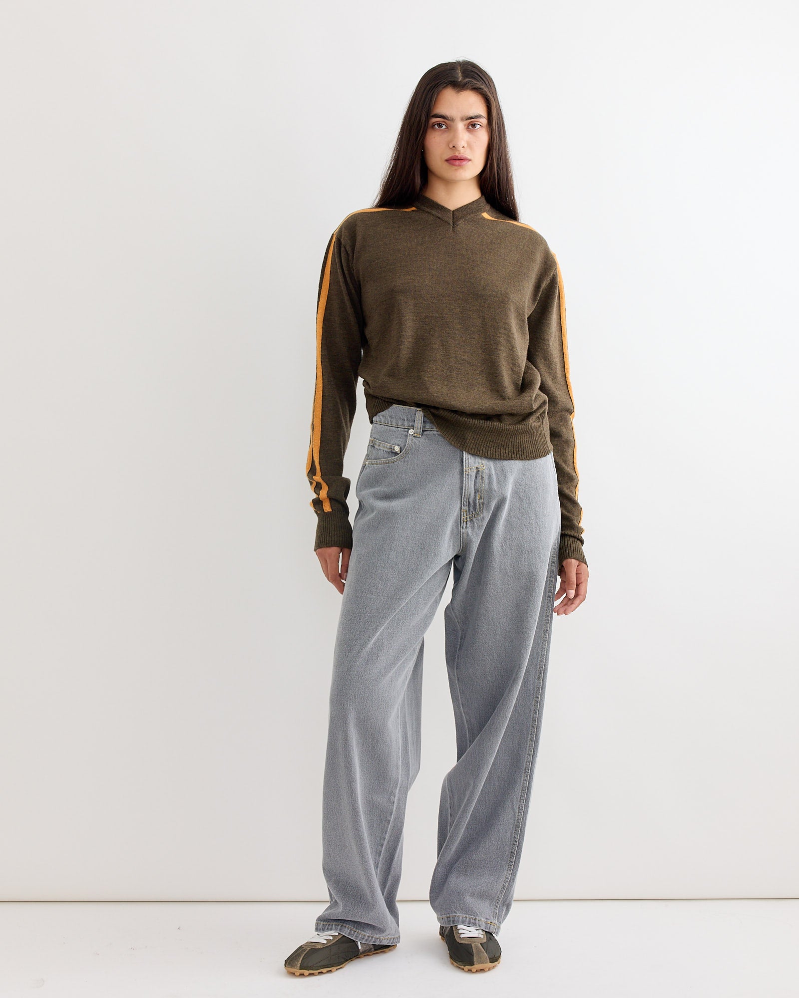 A woman stands against a plain white wall wearing the Gimaguas Baggy Trouser in Grey and a brown long-sleeve sweater with orange stripes. She pairs the look with sneakers and gazes forward with a neutral expression.