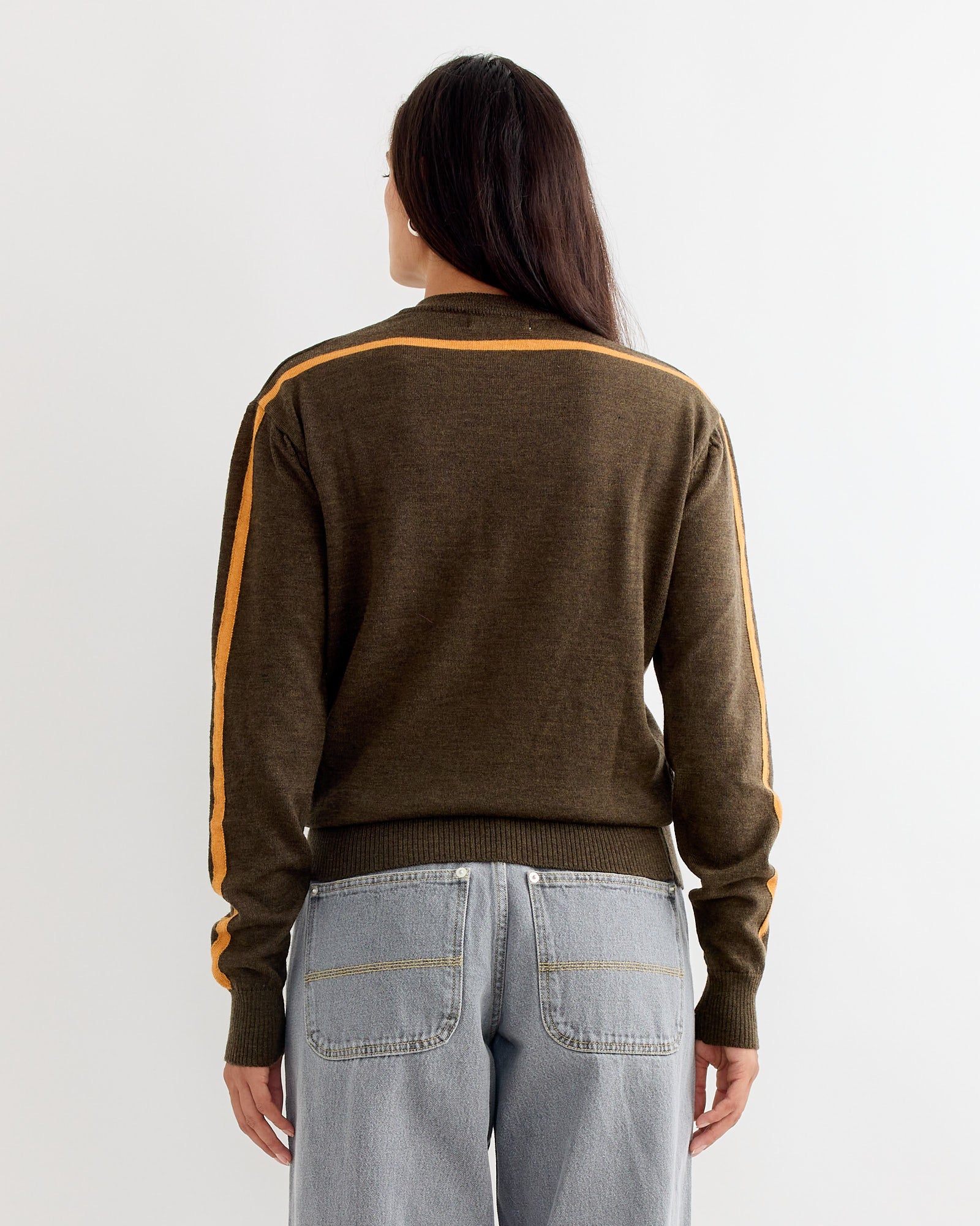 A person with long dark hair, facing away, wears the Gimaguas Sport Jumper in Brown featuring yellow striped sleeves, paired with light blue jeans, all set against a plain light background.