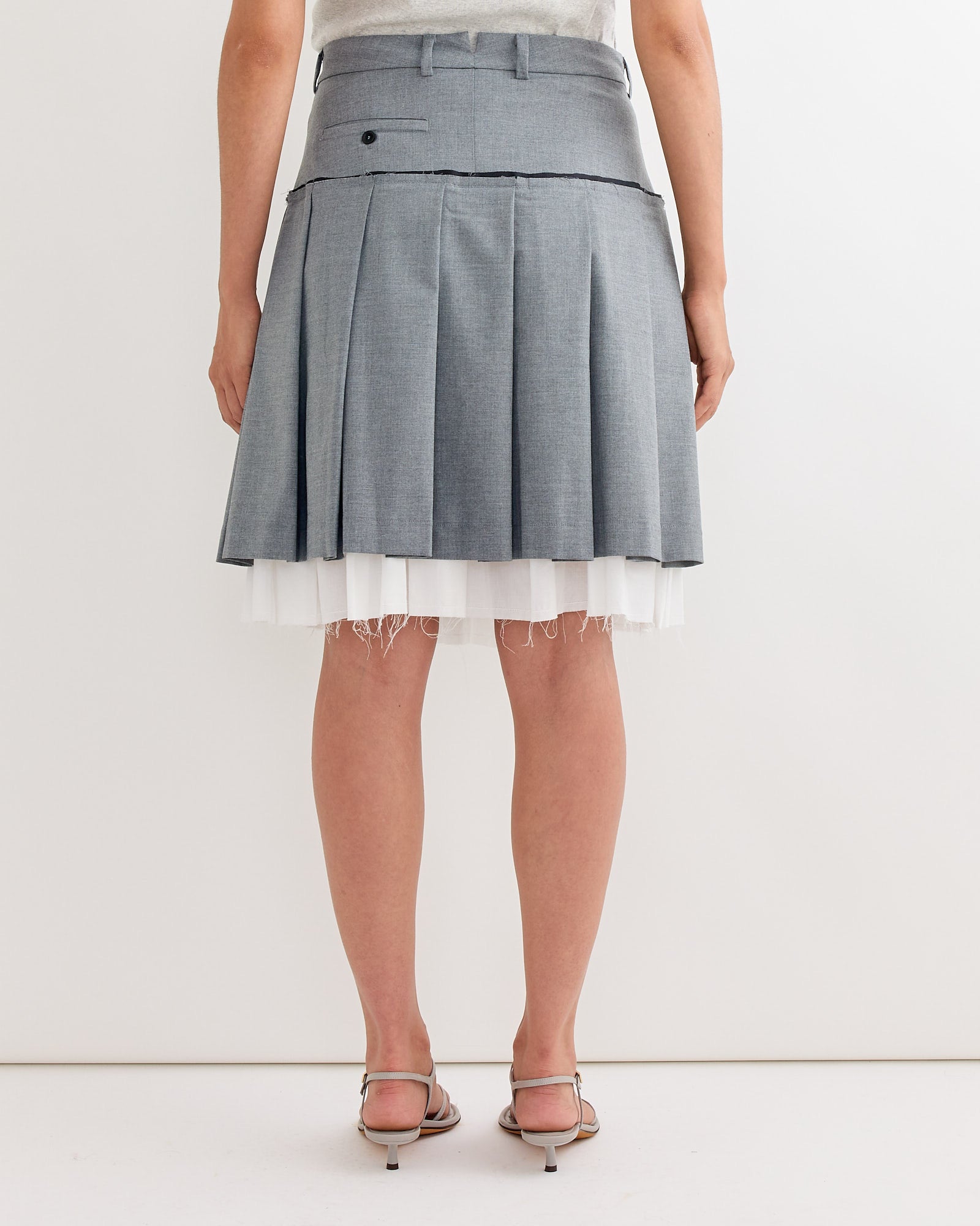 A model faces away, wearing the Gimaguas Tailored Midi Skirt in Grey, featuring pleats and a white tulle underlayer with open-toe heeled sandals, set against a plain white background.