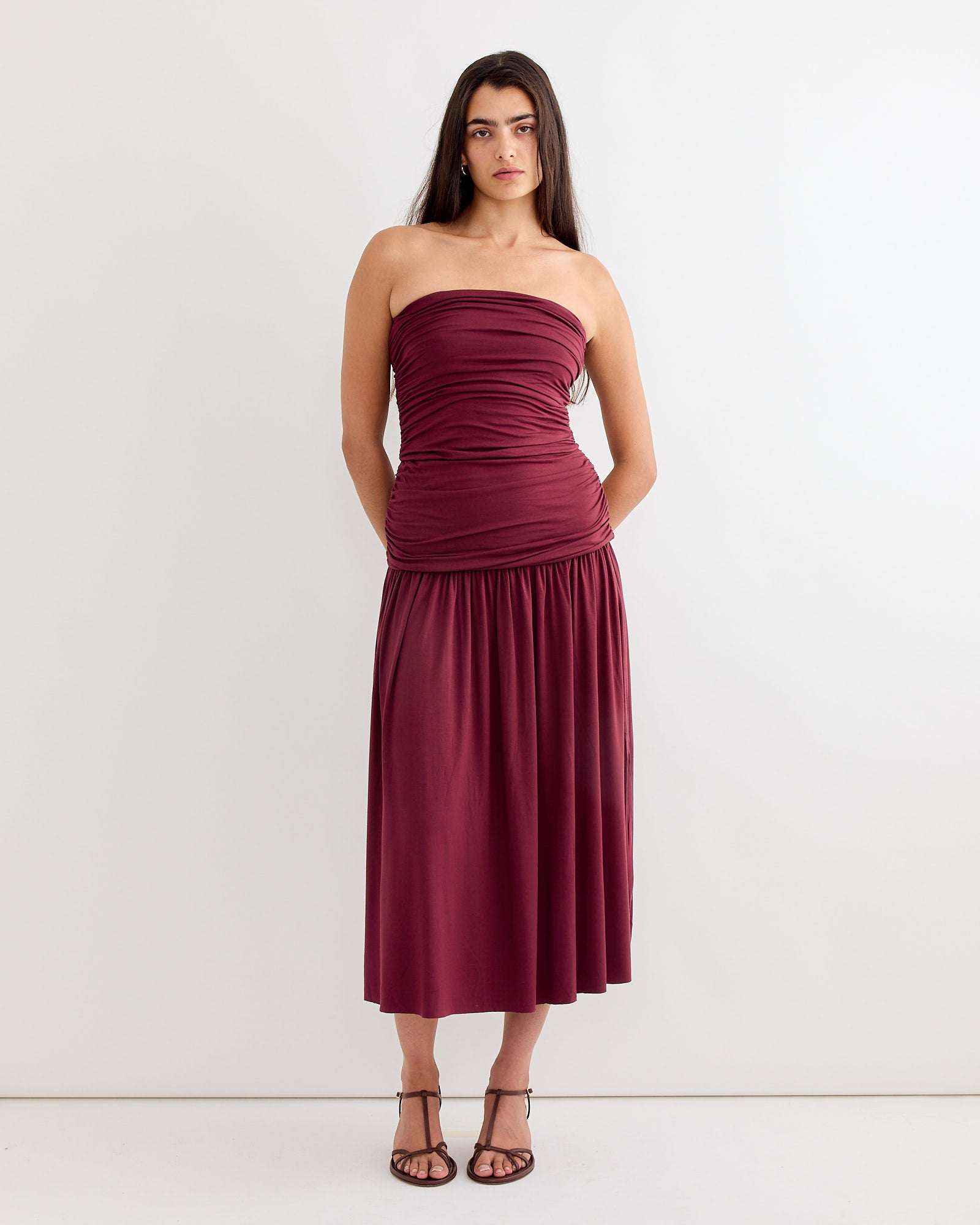 A woman with long, straight dark hair stands against a white background, wearing the Gimaguas Fortuna Maxi Dress in Burgundy, featuring ruched detailing and a calf-length skirt, paired with black strappy heels. Her hands rest at her sides.