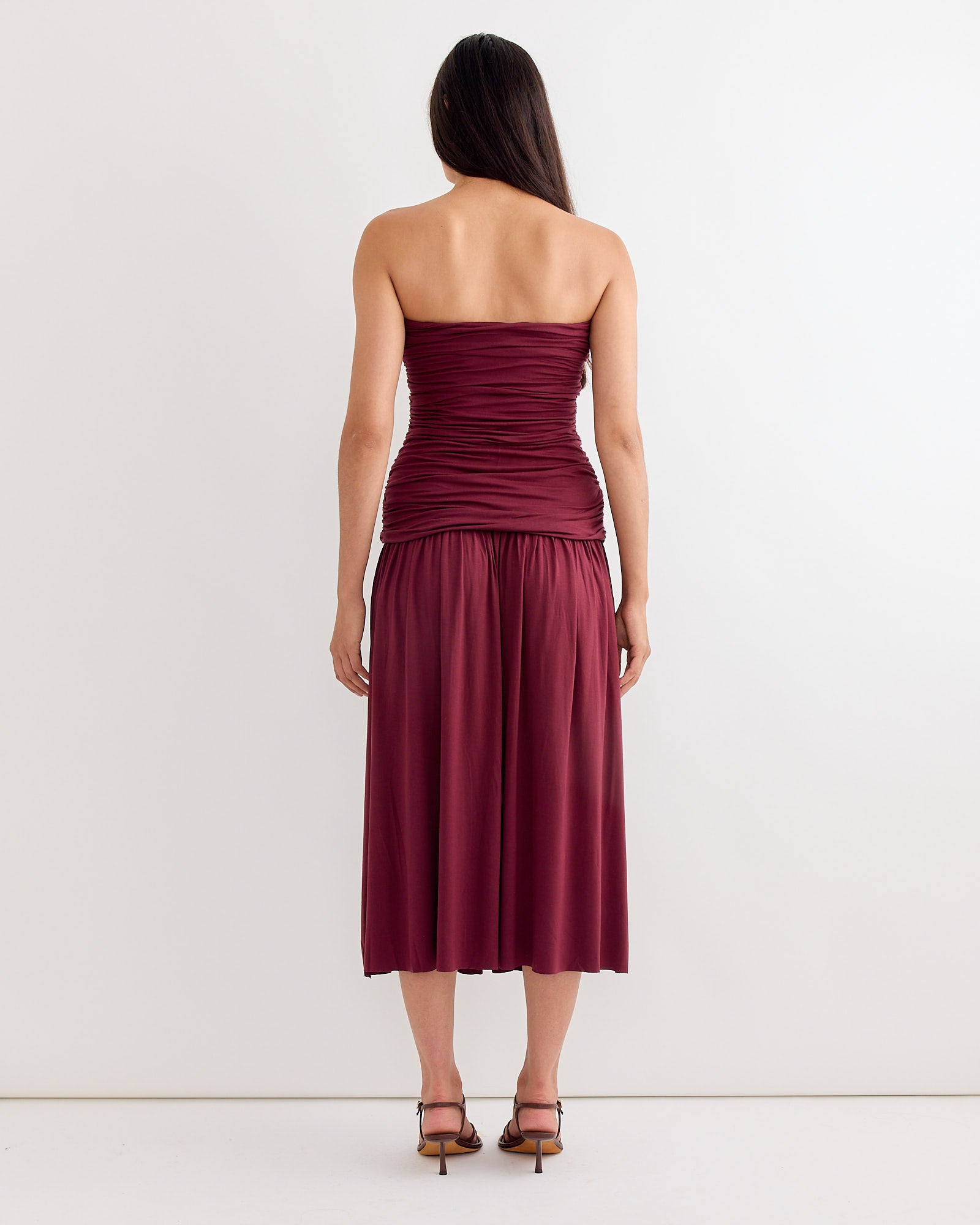 A woman with long dark hair stands facing away, wearing the Gimaguas Fortuna Maxi Dress in Burgundy—strapless with ruching and a flowy, mid-calf skirt—paired with strappy heeled sandals against a plain white background.