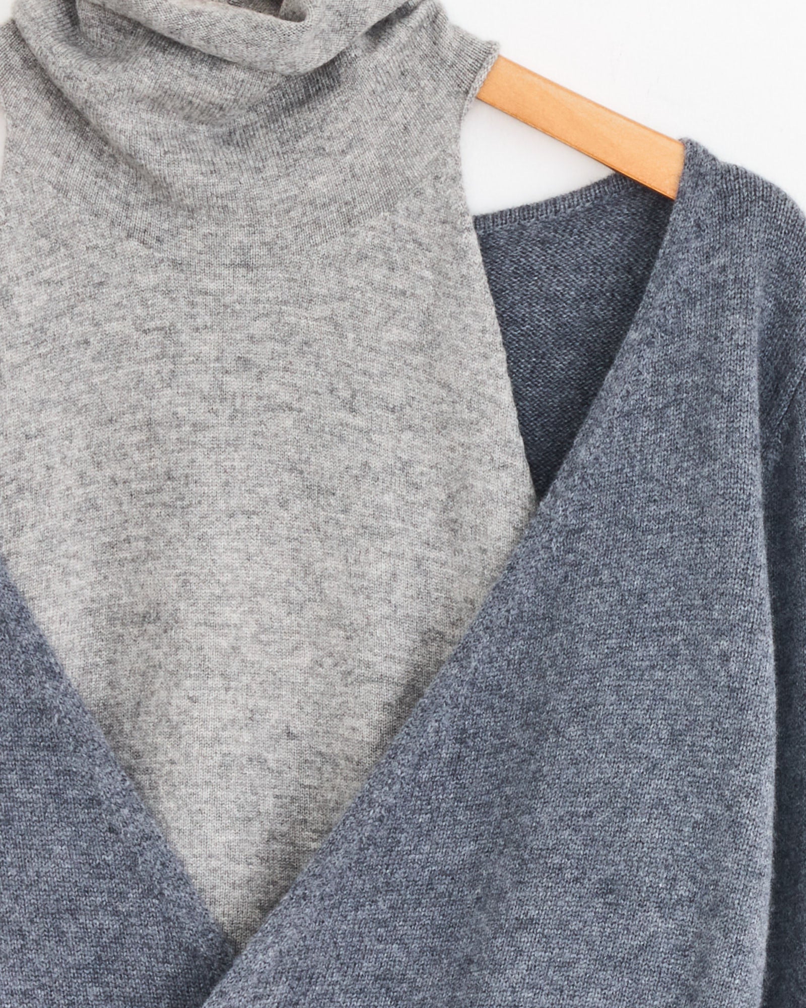 A close-up of the Gimaguas Zante Jumper in Grey, made from recycled fibers, layered under a blue knit sweater—both displayed on a wooden hanger against a white background.