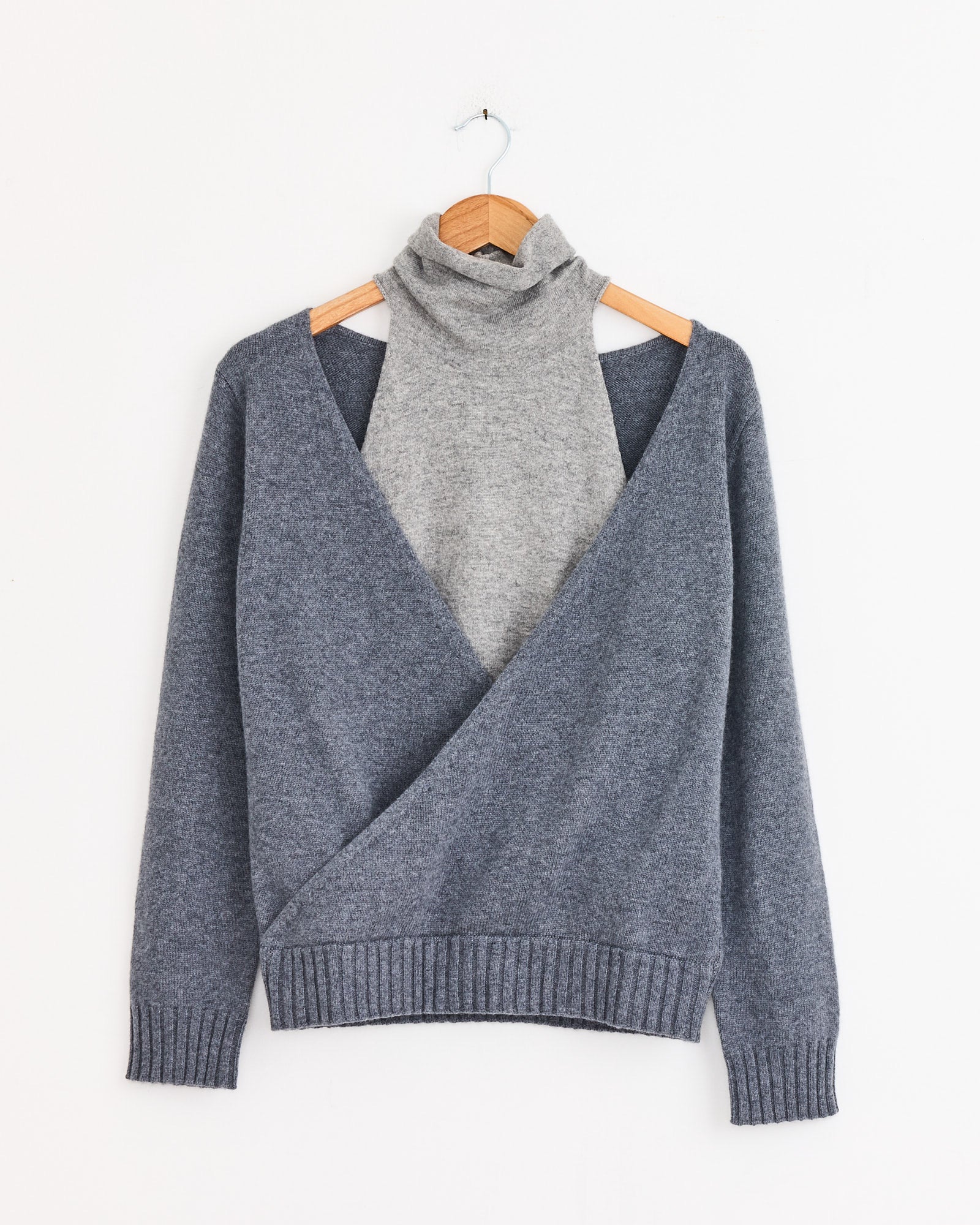 A gray turtleneck sweater layered under the Gimaguas Zante Jumper in Grey, a deep V-neck, long-sleeve knit made from recycled fibers, both displayed on a wooden hanger against a white background.