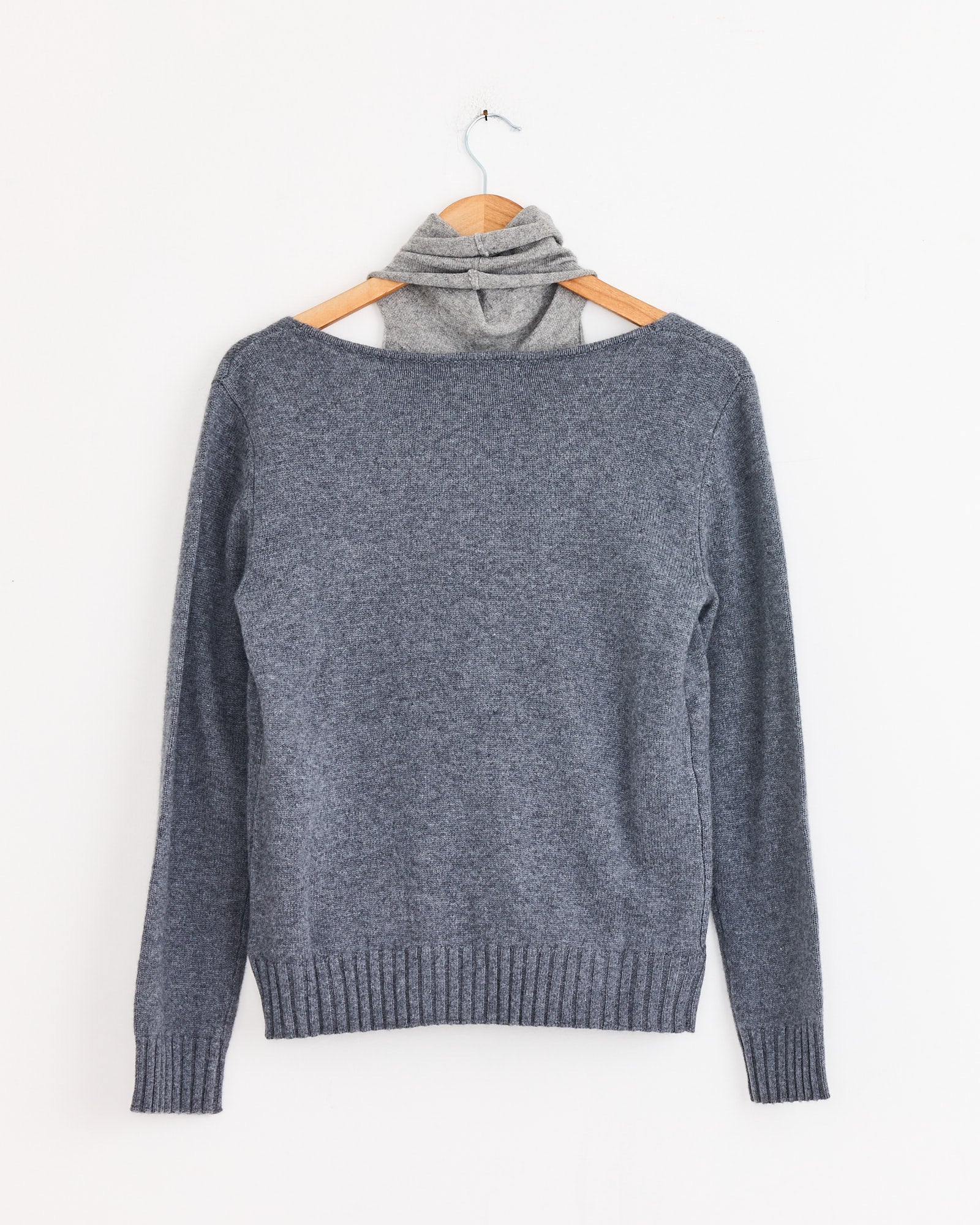 The Zante Jumper in Grey by Gimaguas, made from recycled fibers, hangs on a wooden hanger against a white wall with a light gray turtleneck layered underneath for added style.