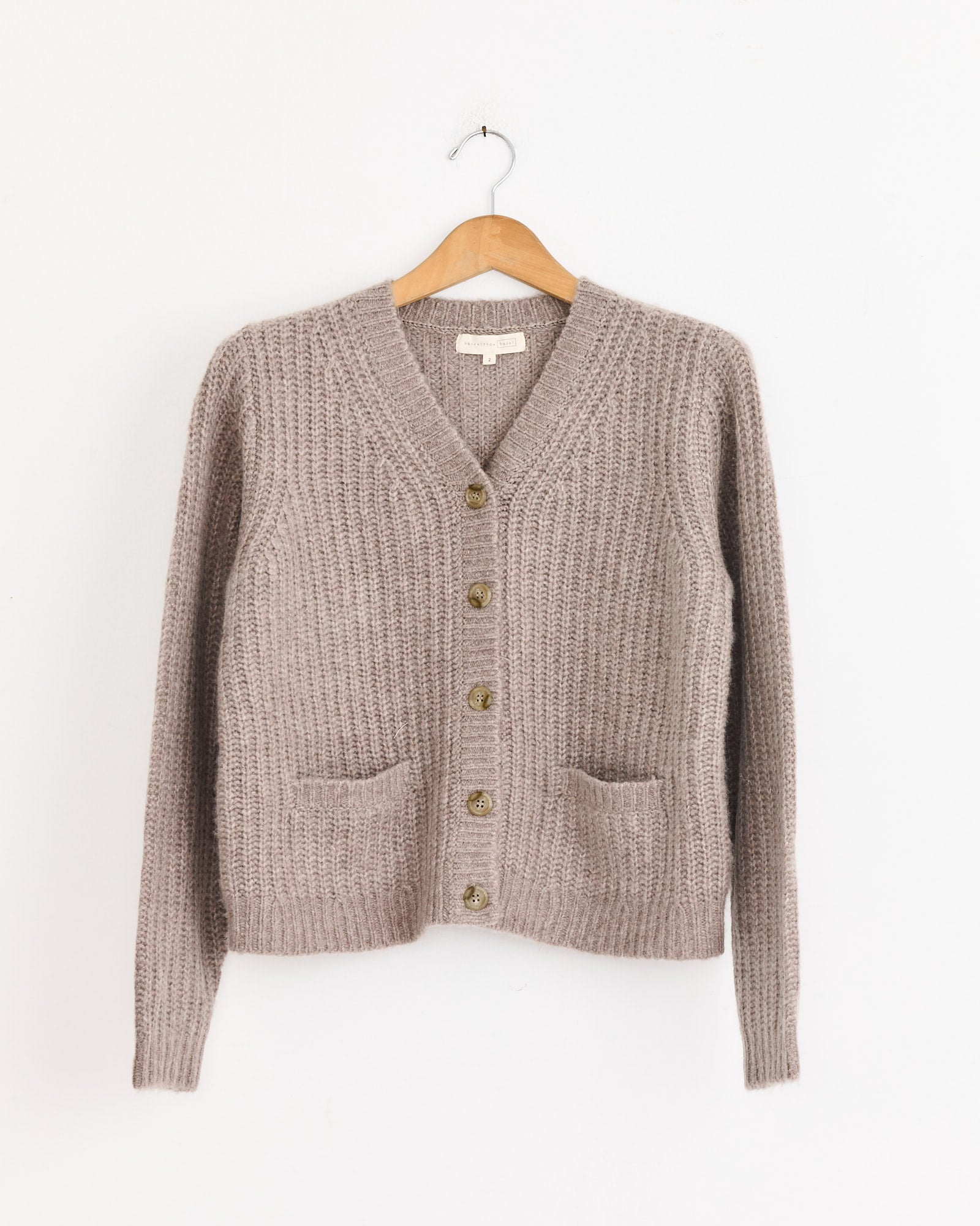 The Hansel From Basel Cashmere Alden Cardigan in Mink features a V-neck, button-down front, two front pockets, and long sleeves, shown on a wooden hanger against a white background.