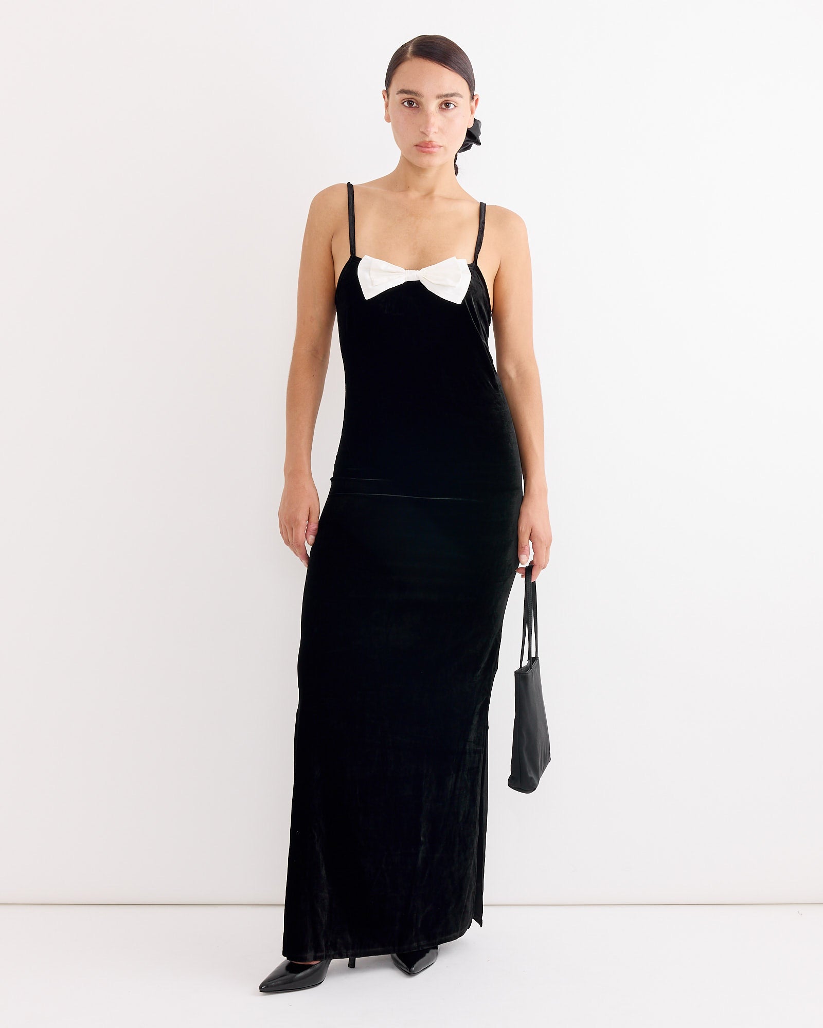 A woman stands against a white background wearing the Hai Antoinette Dress in Black/Ivory, a long sleeveless velvet silk dress with thin straps and a large ivory bow at the chest. She holds a small black bag, wears black shoes, and her hair is styled back.