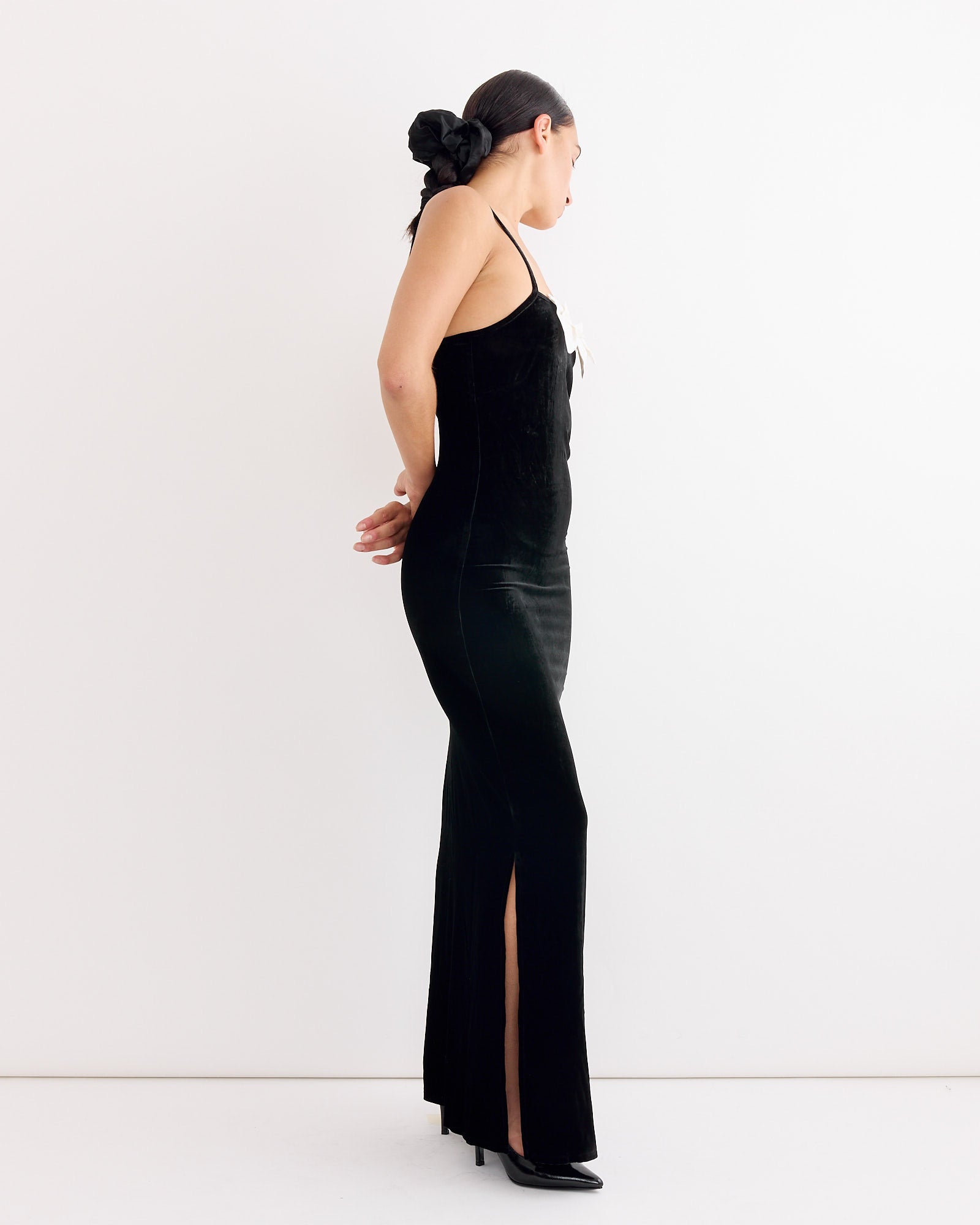 A woman stands sideways against a white background, wearing Hai's Antoinette Dress in Black/Ivory, styled with a large black scrunchie in her tied-back hair and black heels.