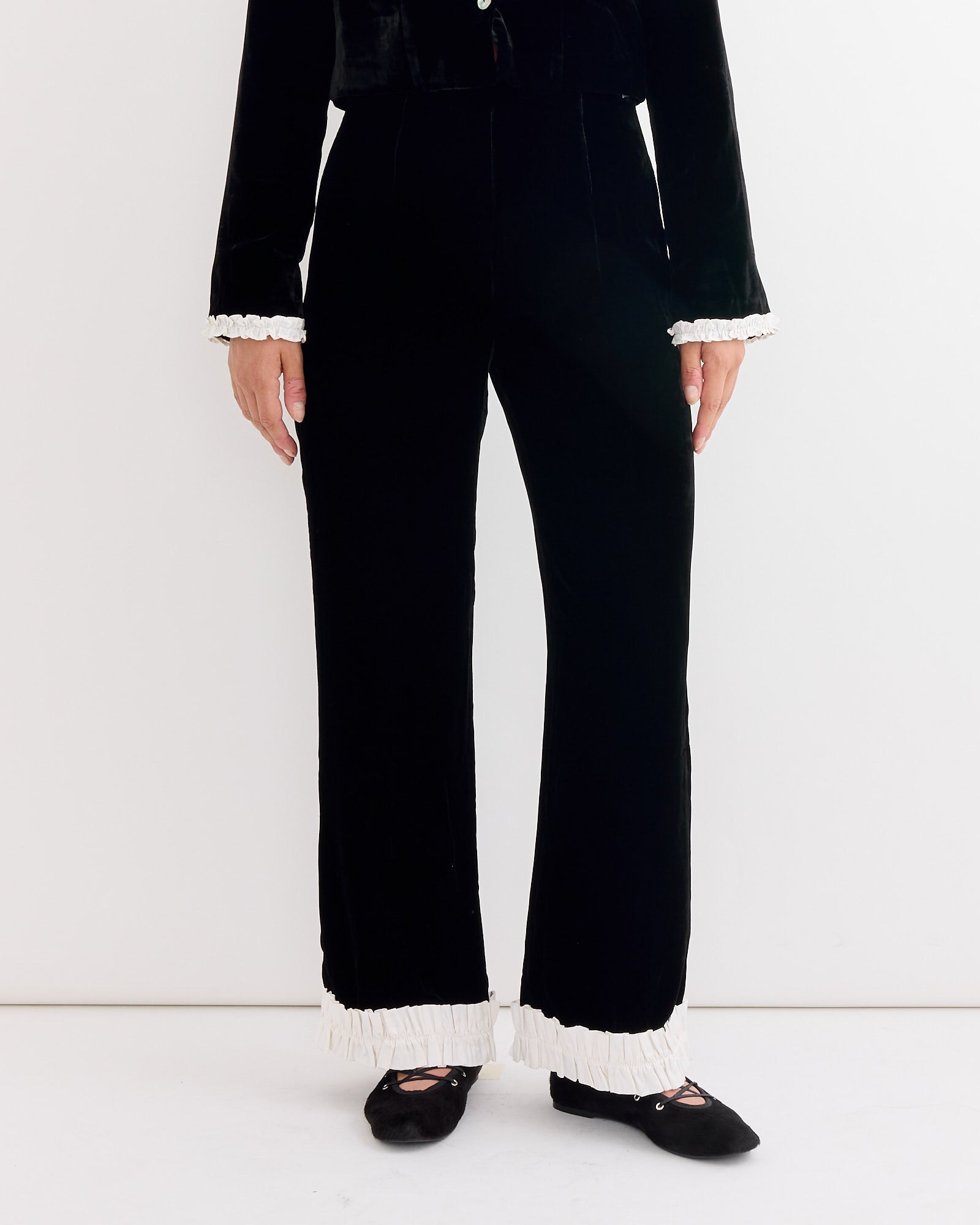 A person stands against a white wall wearing Hai's Leo Velvet Trouser in Black/Ivory, paired with a matching black top featuring white ruffled sleeves, and black flat shoes.