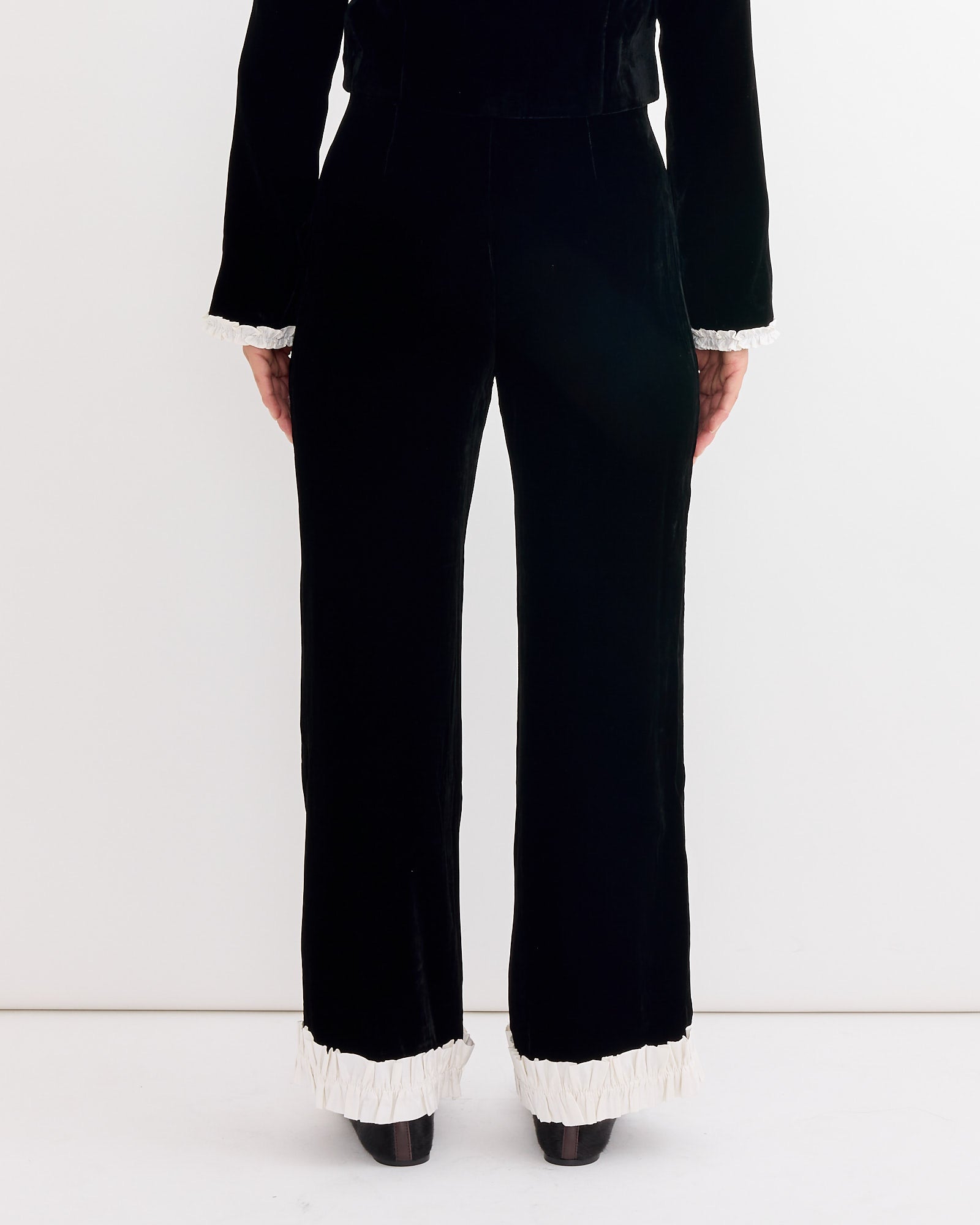 A person stands facing away, showing only the lower torso and legs, wearing Hai's Leo Velvet Trouser in Black/Ivory with a high-rise waist, subtly flared legs, and white silk ruffles at the ankles, set against a plain white background.