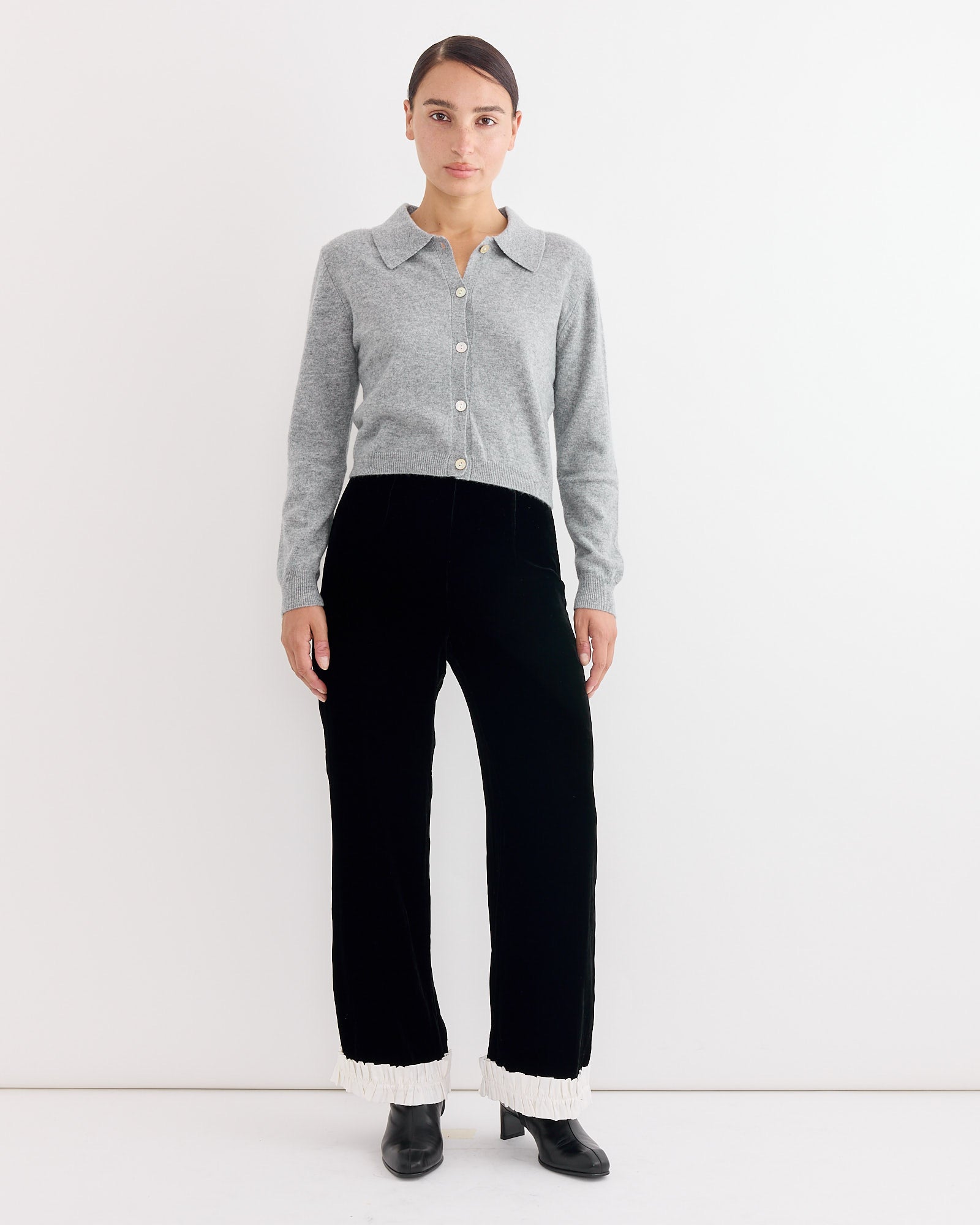 A woman with dark hair tied back and a neutral expression wears the Hai Anselma Cardigan in Grey, paired with high-waisted black velvet pants featuring white ruffled cuffs and black ankle boots, standing against a white background.