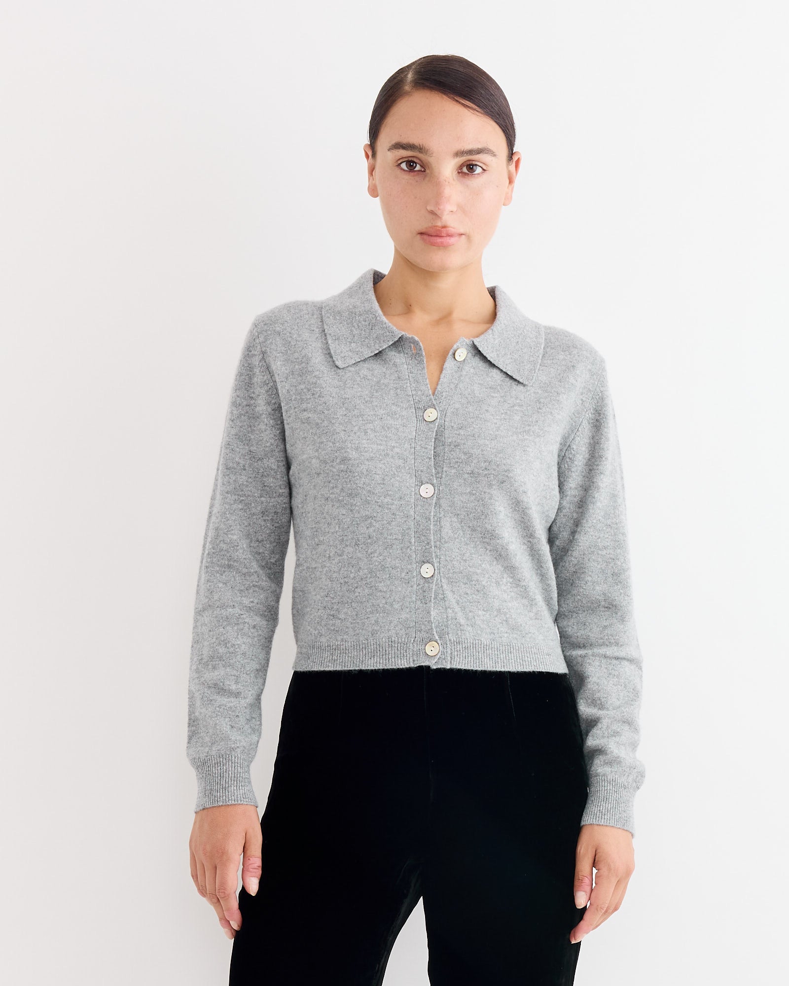 A woman with dark hair tied back wears the Hai Anselma Cardigan in Grey, a slim fit, collared style, paired with black pants. She stands facing forward with a neutral expression against a plain white background.
