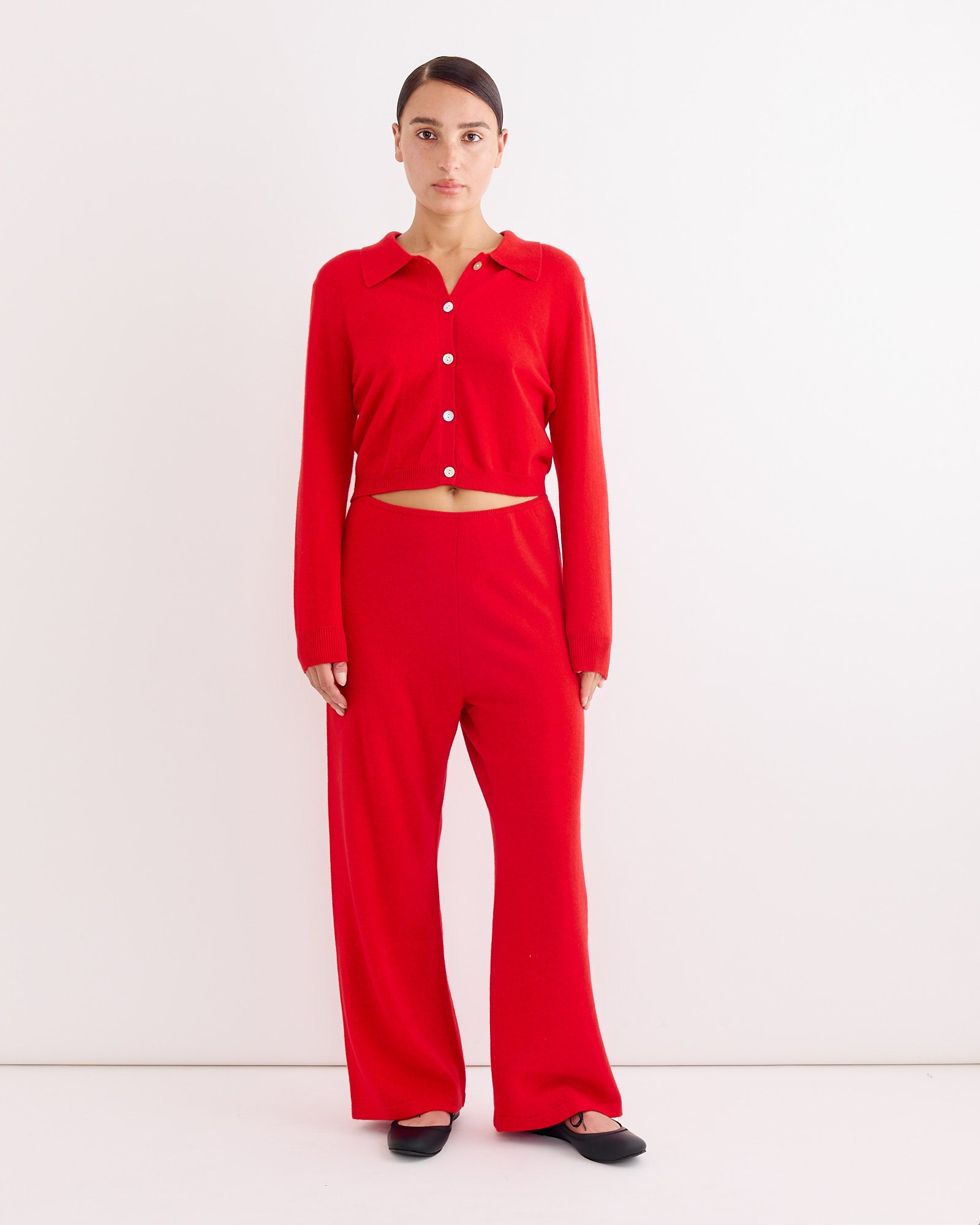 A woman stands against a white wall wearing the Hai Anselma Cardigan in Red, paired with matching wide-leg pants. Her straight brown hair is pulled back and she completes the look with black flats.