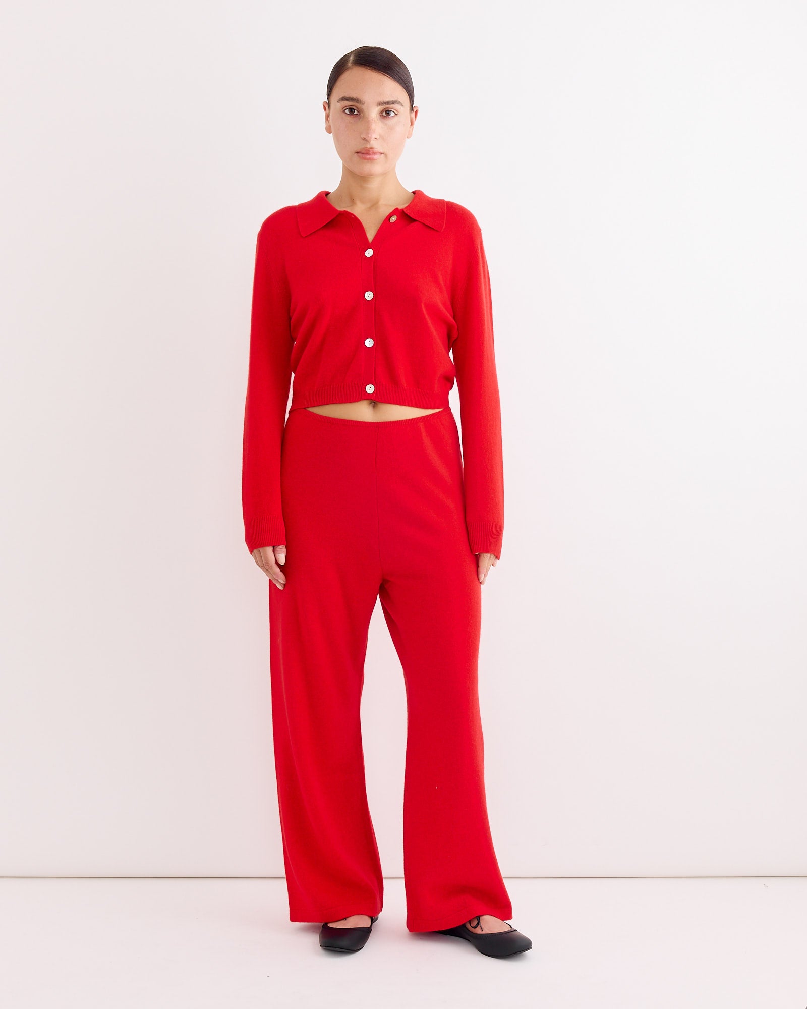 A woman stands facing forward against a plain white background, wearing Hai’s Alfred Trouser in Red with a matching red long-sleeve button-up top. Her hair is pulled back and she’s wearing black shoes.