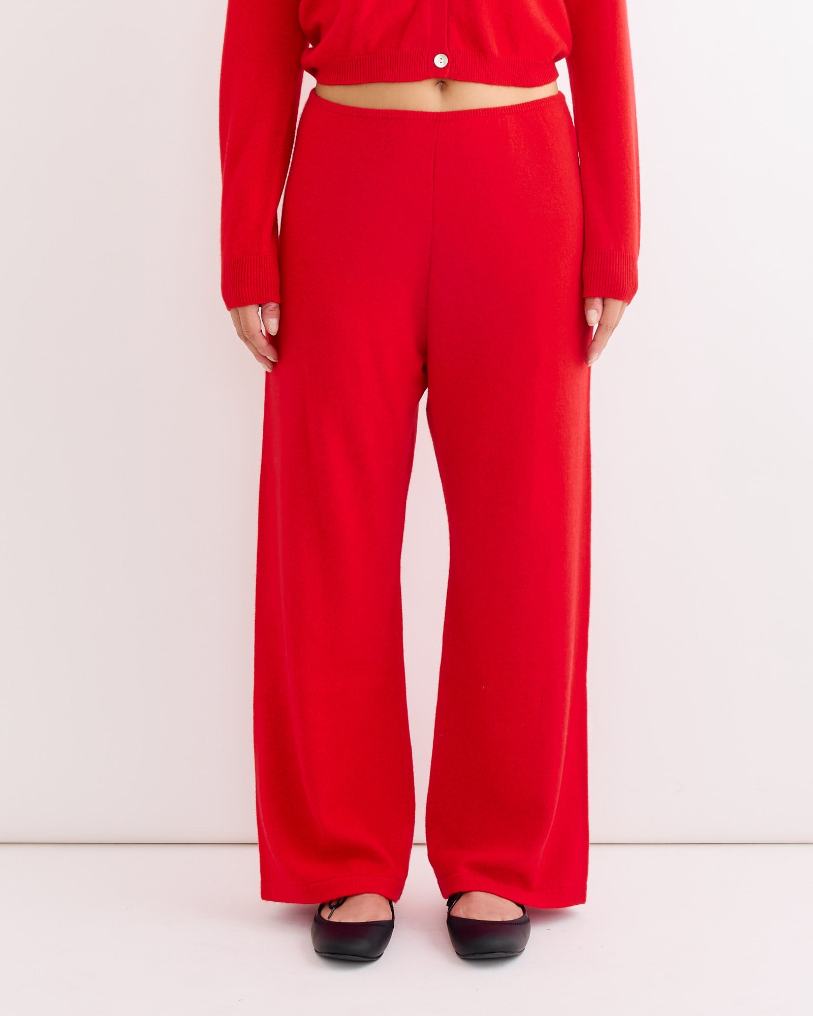 Against a white background, a person wears Hai’s Alfred Trouser in Red paired with a matching red long-sleeve buttoned top and black ballet flats, standing with hands relaxed at their sides.
