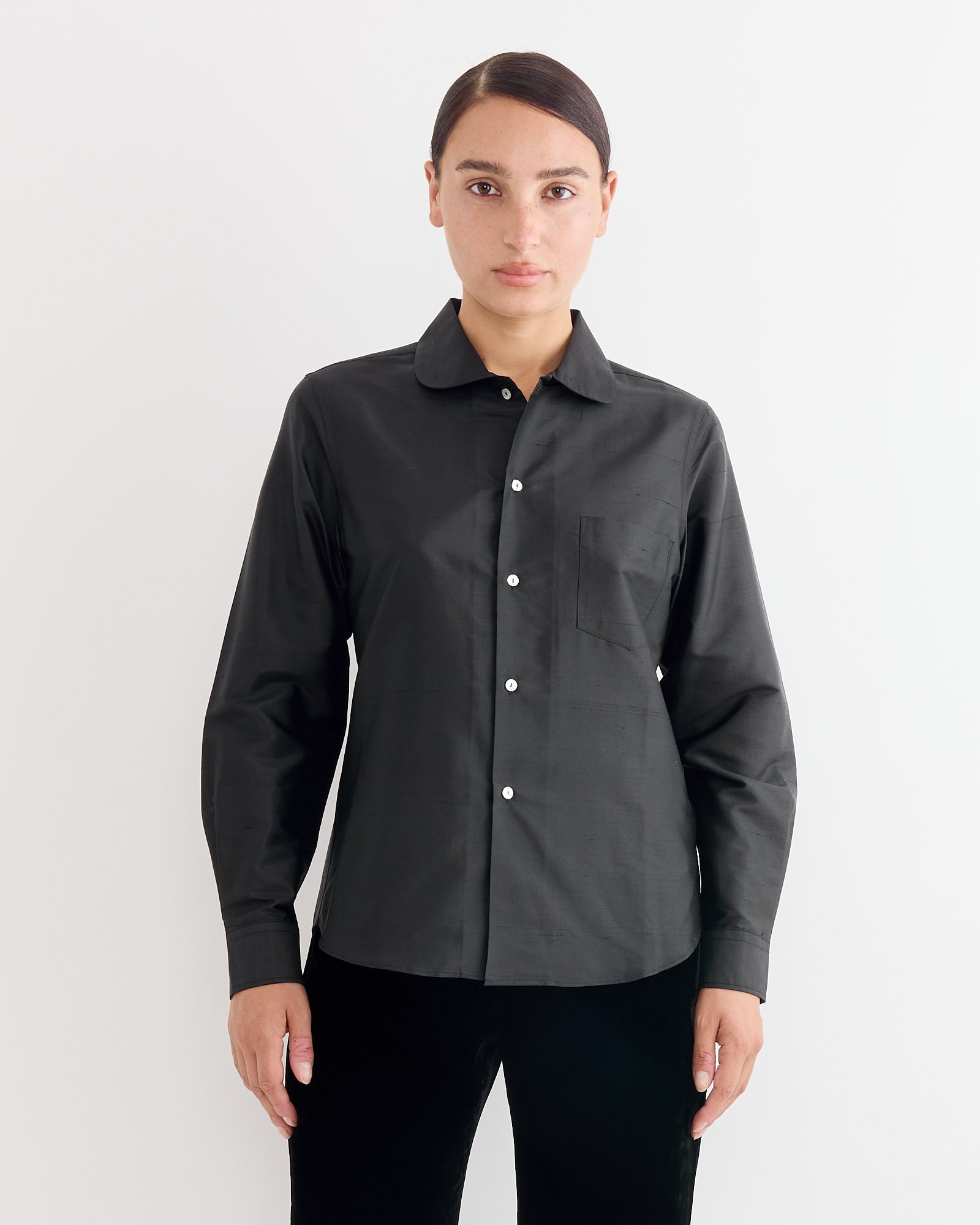 A person with dark hair pulled back wears the Hai Elia Shirt in Black, a long-sleeve silk top with a chest pocket, standing against a plain white background.
