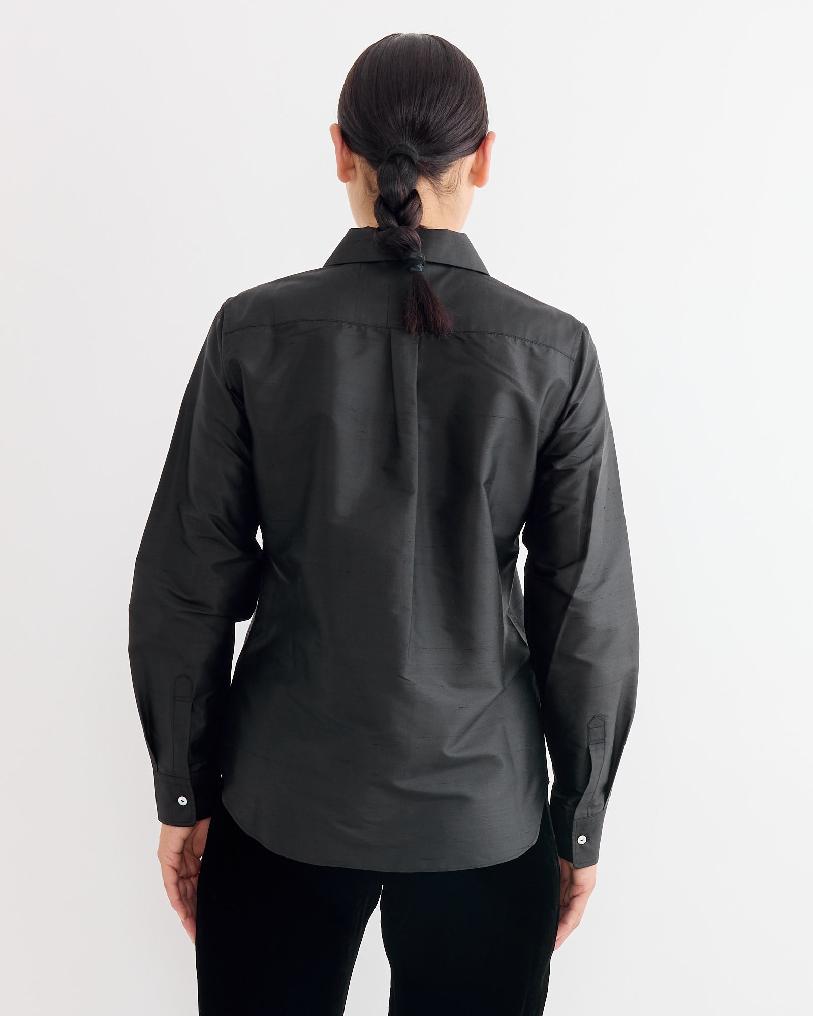 A person with dark braided hair is shown from behind against a white background, wearing the Hai Elia Shirt in Black and black pants.