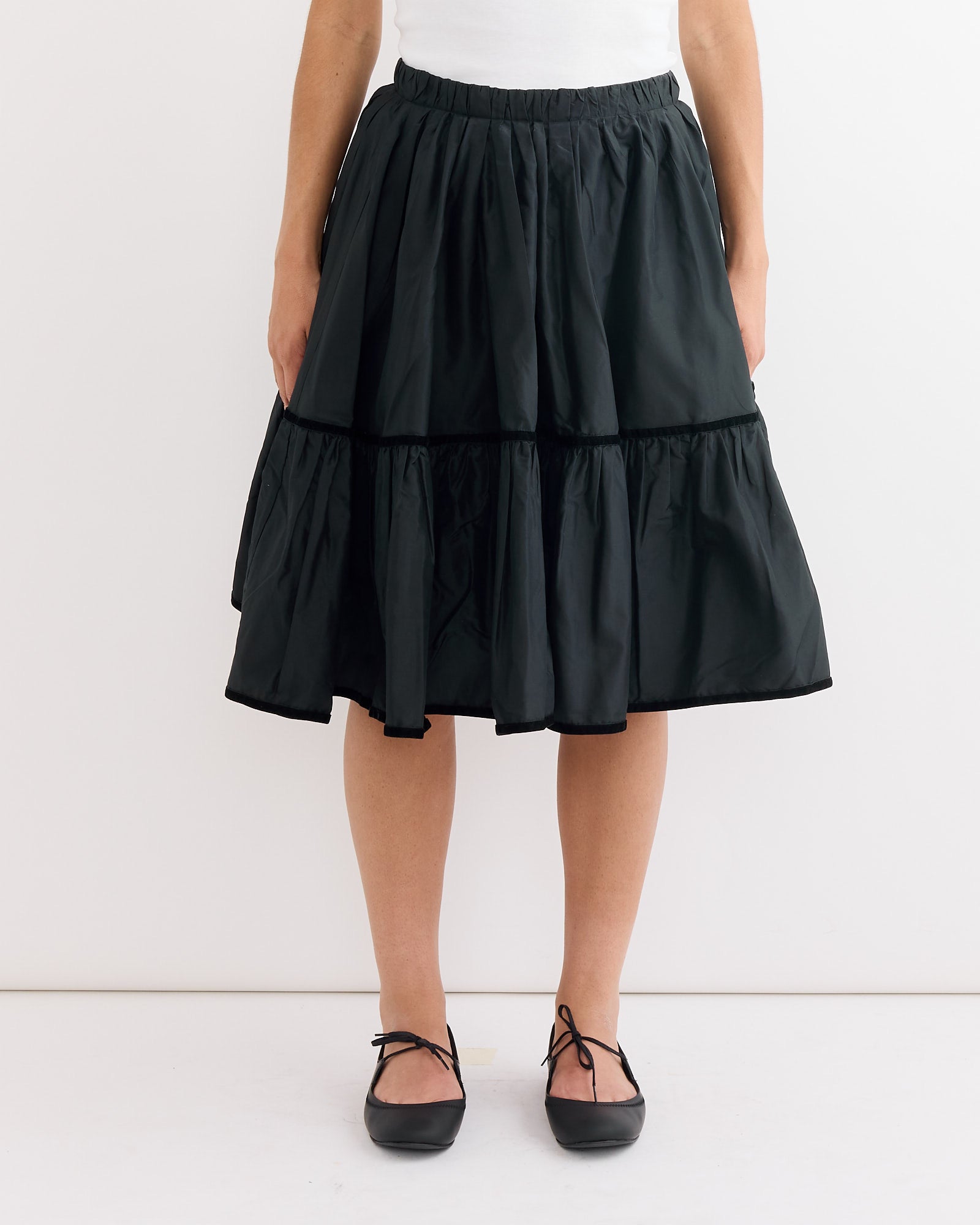 A woman wearing the Hai Ada Skirt in Black—a knee-length taffeta silk skirt with a gathered waistband—paired with black ballet flats, stands against a plain white background.