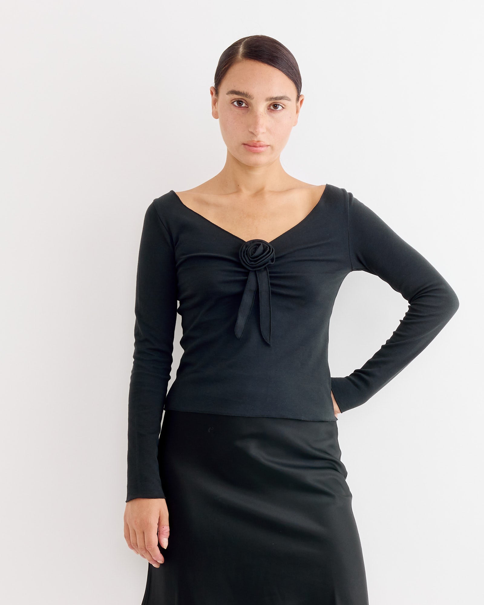 A woman with dark hair tied back wears the Hai Gretchen Tee in Black—an organic cotton jersey long-sleeve v-neck with a central rosette—paired with a black satin skirt, standing against a plain white background.